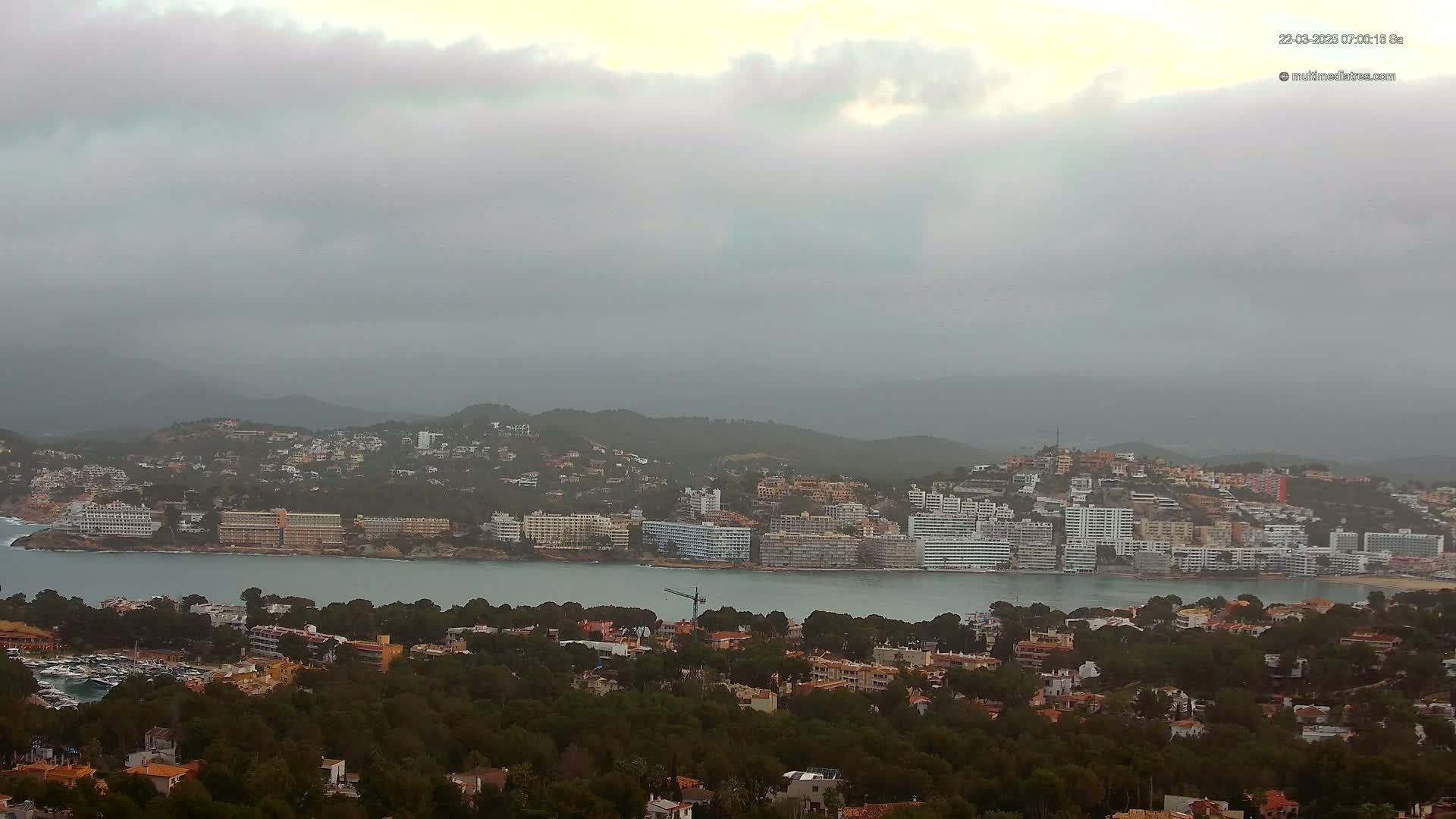 A coastal town nestled among hills is shrouded in an overcast sky, with a calm body of water separating the town from a densely wooded area.