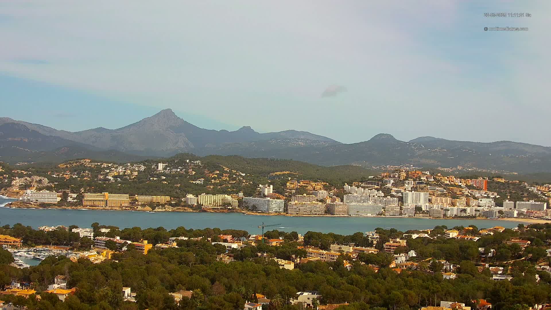 A coastal town nestled among green hills and mountains under a mostly clear sky, with a calm body of water separating the town from a shoreline dotted with hotels and buildings.