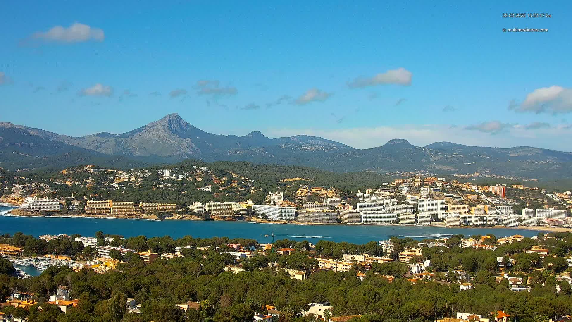A coastal town nestled amongst green hills and mountains under a clear blue sky with a few scattered clouds.