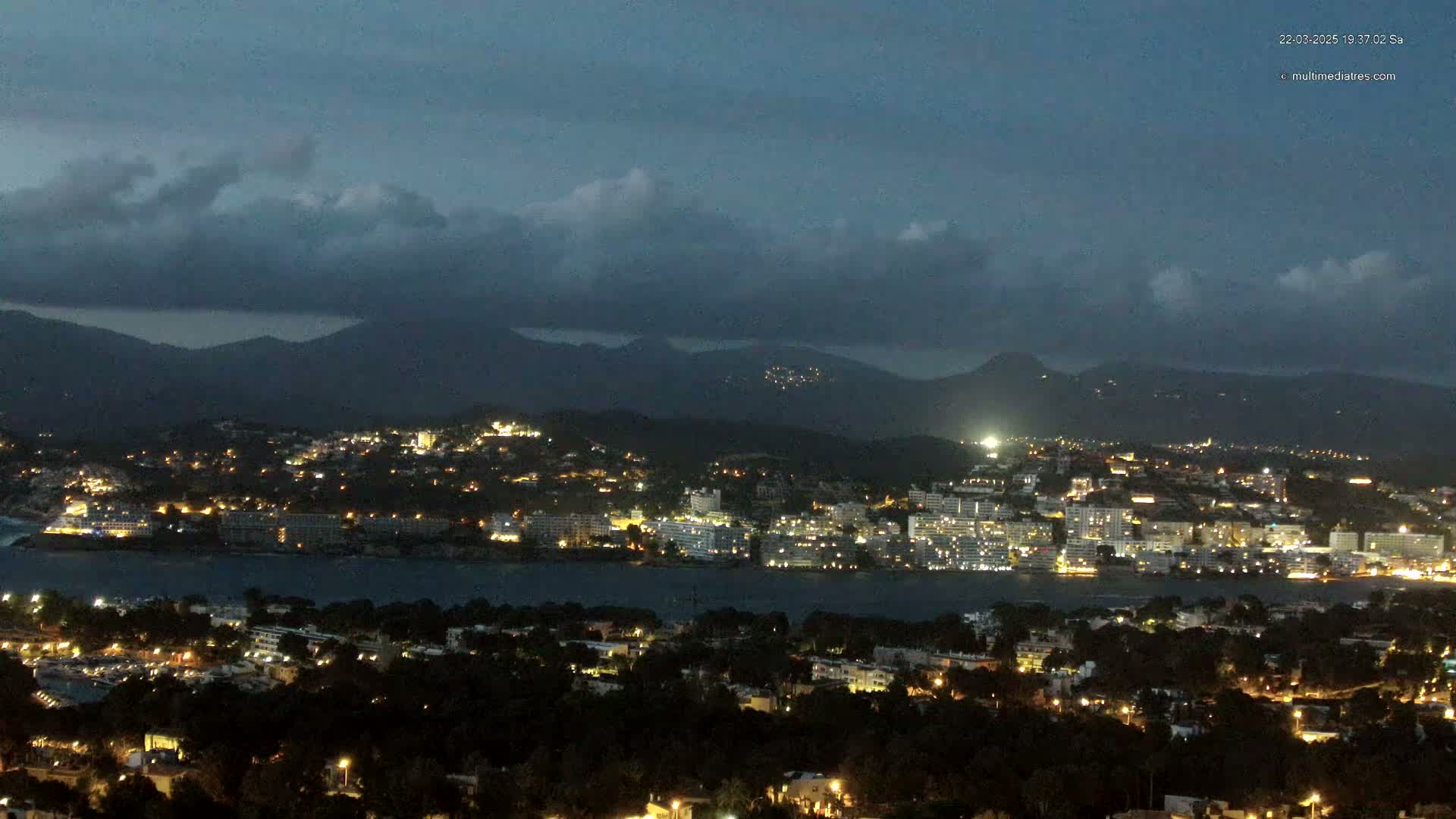 A nighttime view of a coastal town illuminated with lights, situated beneath a cloudy, dark sky and backed by dark mountains.