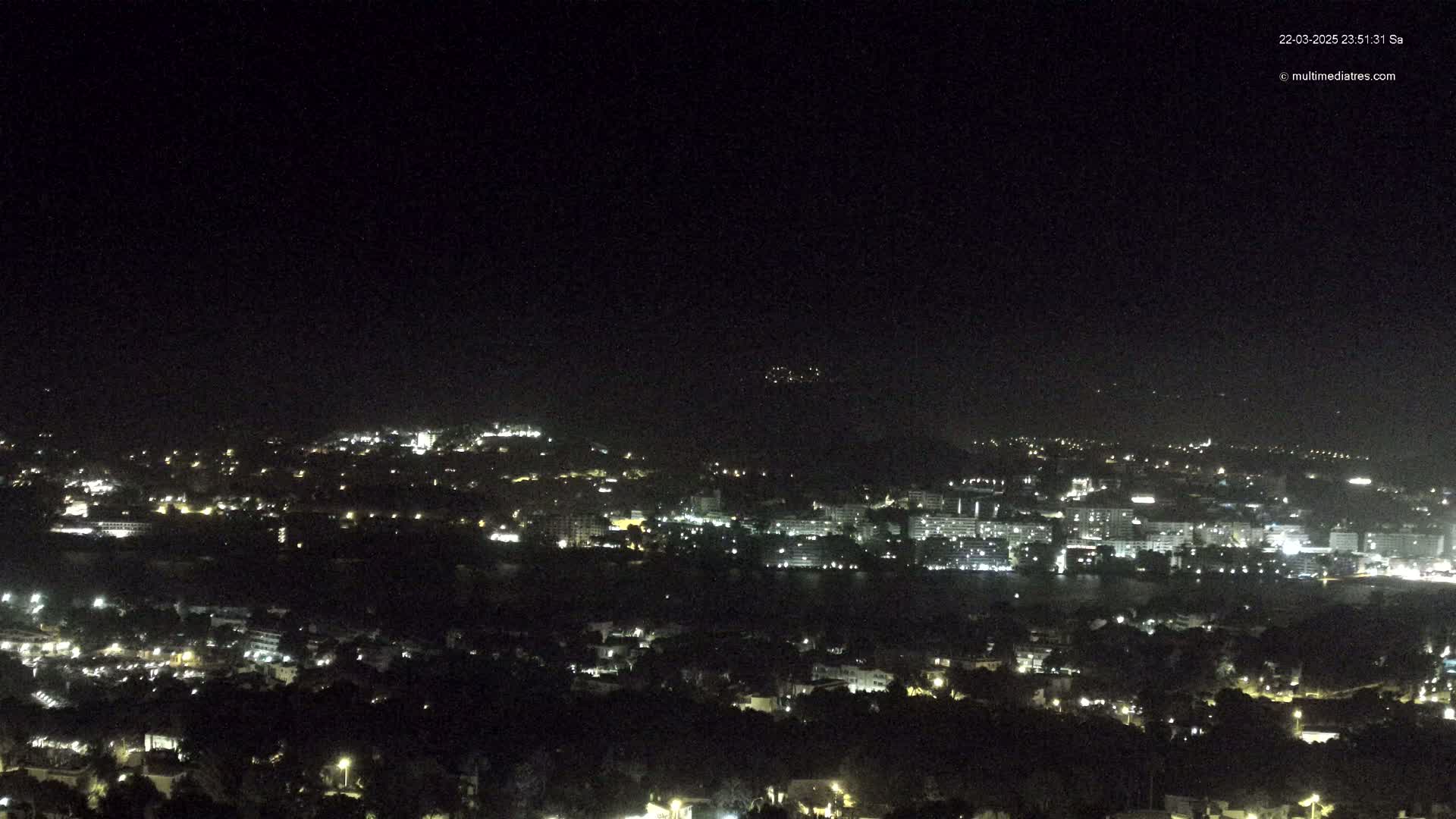 A nighttime view of a coastal city illuminated by scattered lights, under a clear, dark sky.