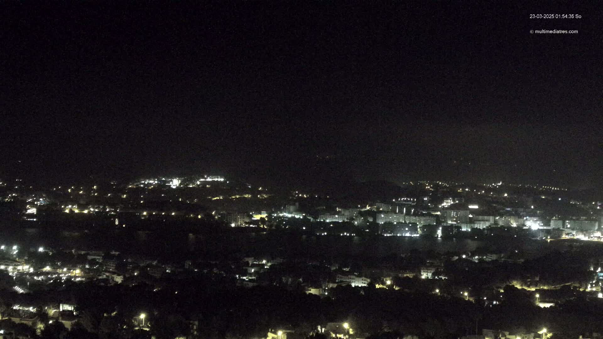 A nighttime, long-range view of a coastal city illuminated with scattered lights under a dark, clear sky.