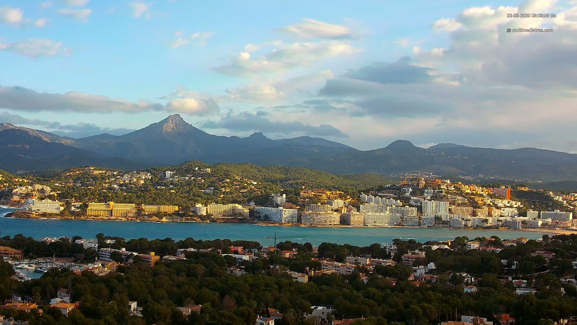 A coastal town nestled between a calm bay and a mountain range under a partly cloudy sky.