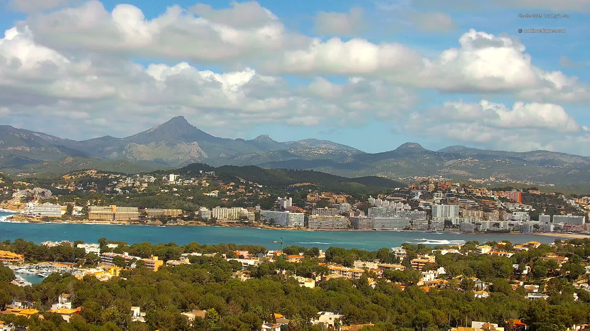 A coastal town nestled amongst lush greenery and backed by a range of mountains under a partly cloudy sky.