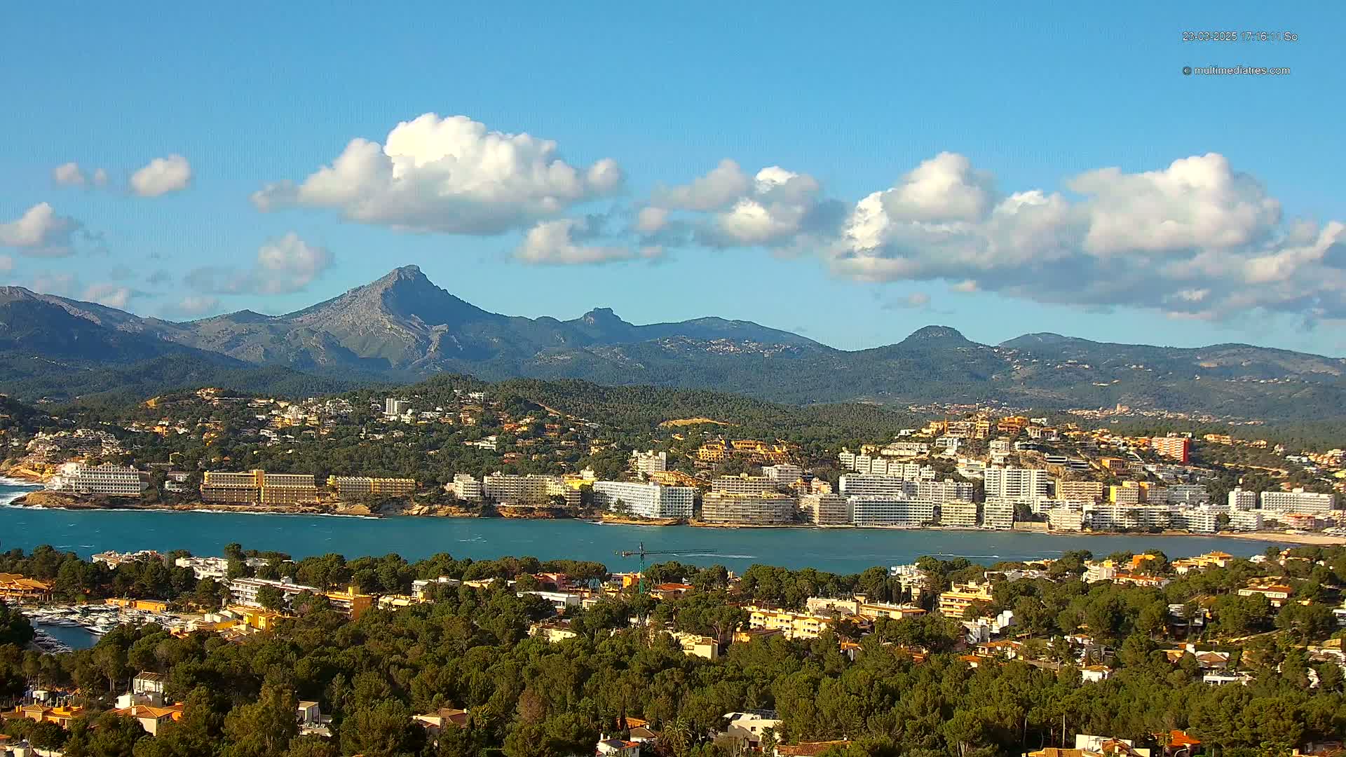 A coastal town nestled between a calm bay and a mountain range under a mostly sunny sky with scattered cumulus clouds.
