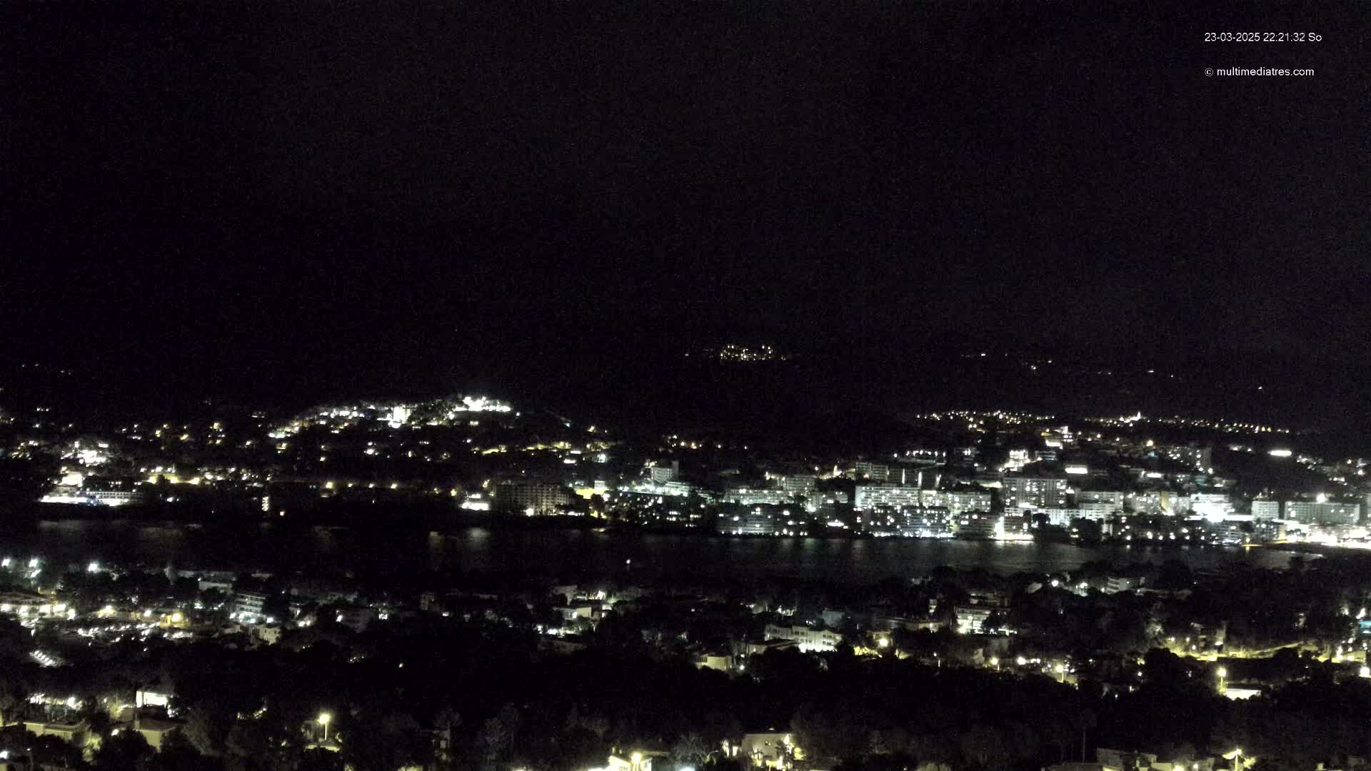 A nighttime, high-angle view shows a coastal city with numerous lights illuminating buildings and streets, and a dark body of water separating a section of the city from another.