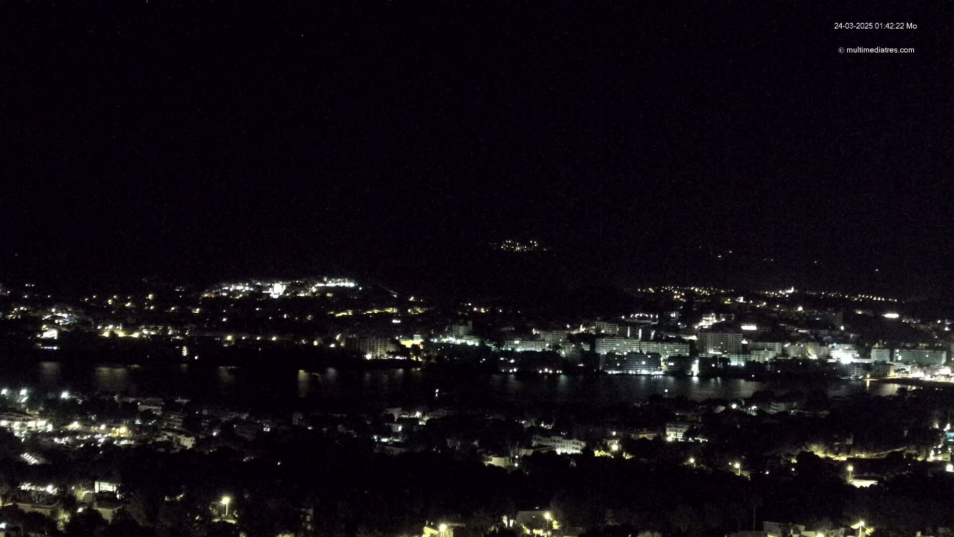 A nighttime, high-angle view of a coastal city with numerous lights illuminating buildings and streets,  partially surrounded by dark water.