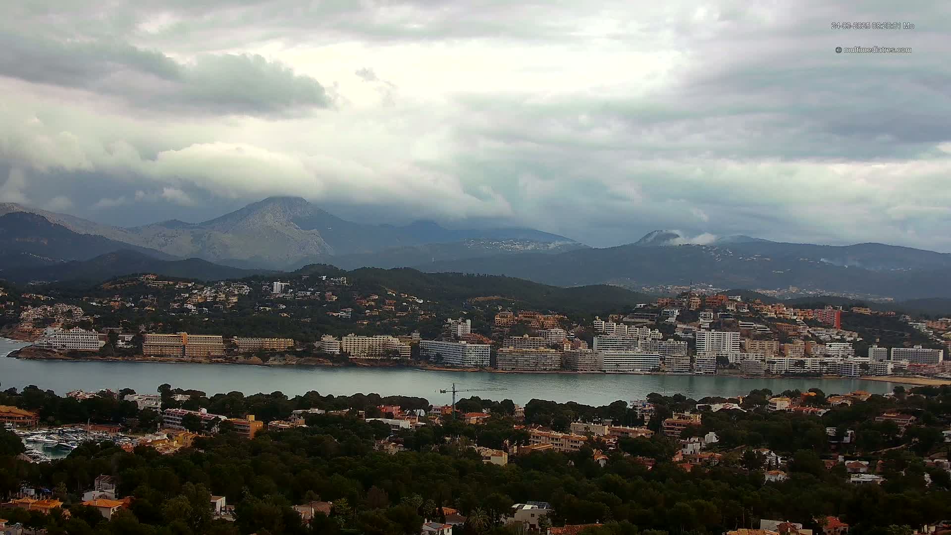 A coastal town nestled among green hills and mountains under a cloudy sky sits beside a calm body of water.