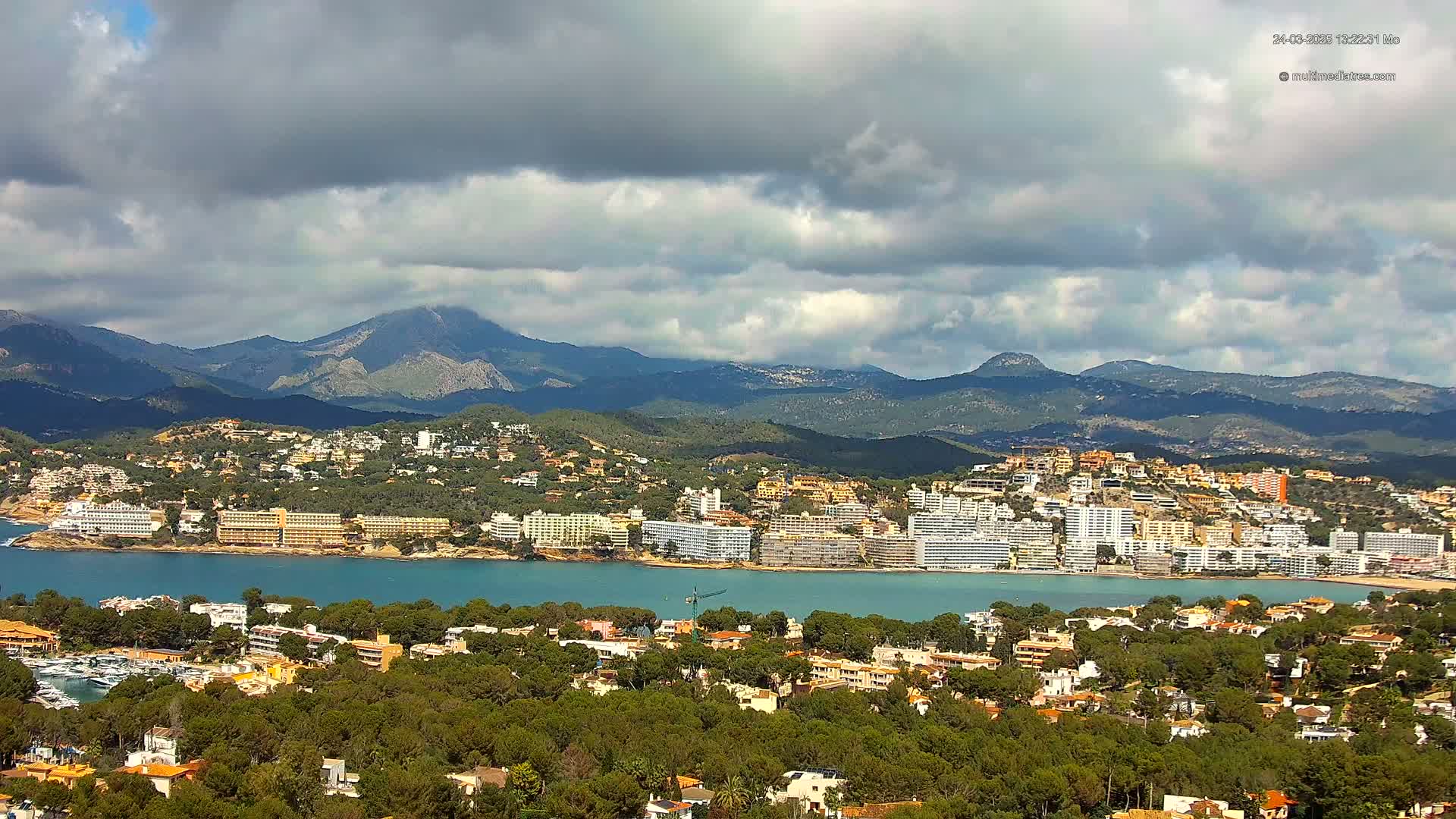 A coastal town nestled between a calm bay and a mountainous backdrop under a partly cloudy sky.