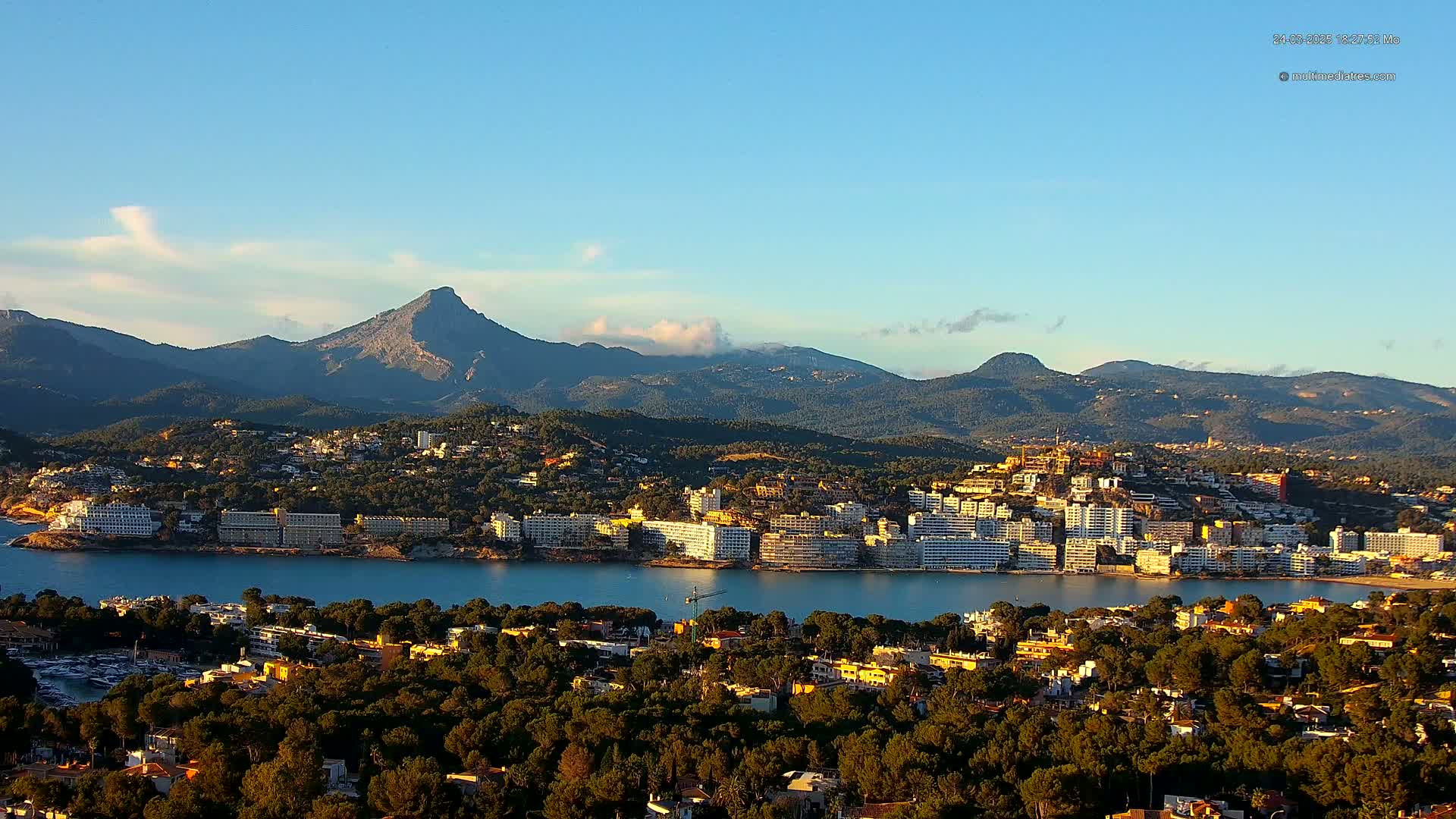 A coastal town nestled between a calm bay and a mountain range under a clear blue sky.