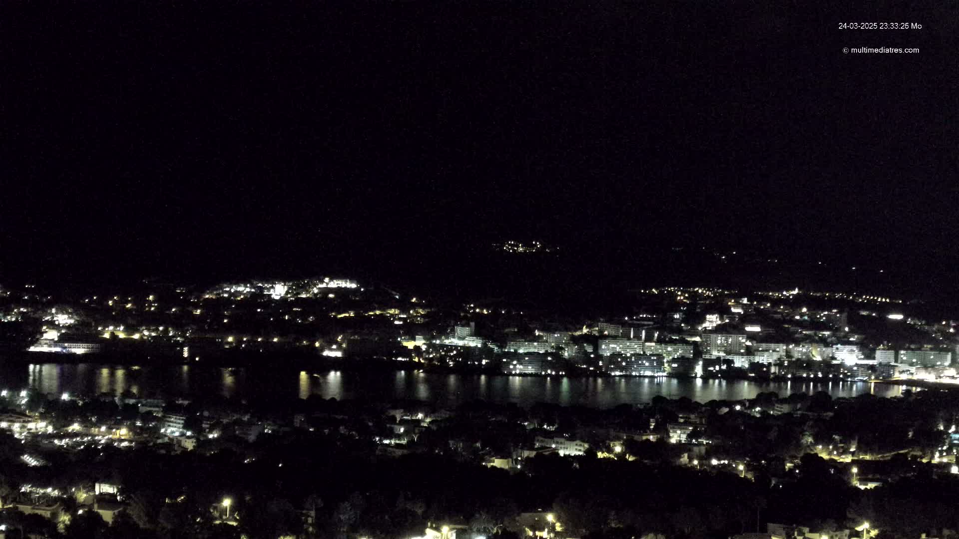 A nighttime, aerial view shows a city with lights illuminated along a body of water under a clear, dark sky.