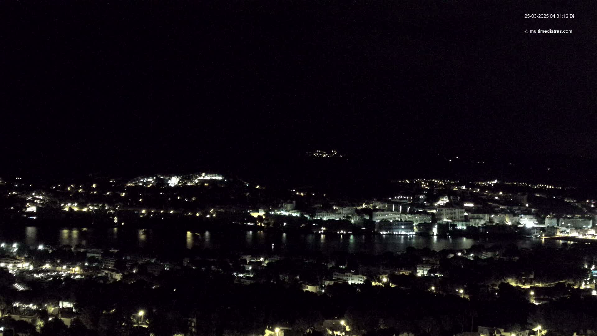 A nighttime, high-angle view of a city with lights illuminating buildings and a body of water, under a clear, dark sky.