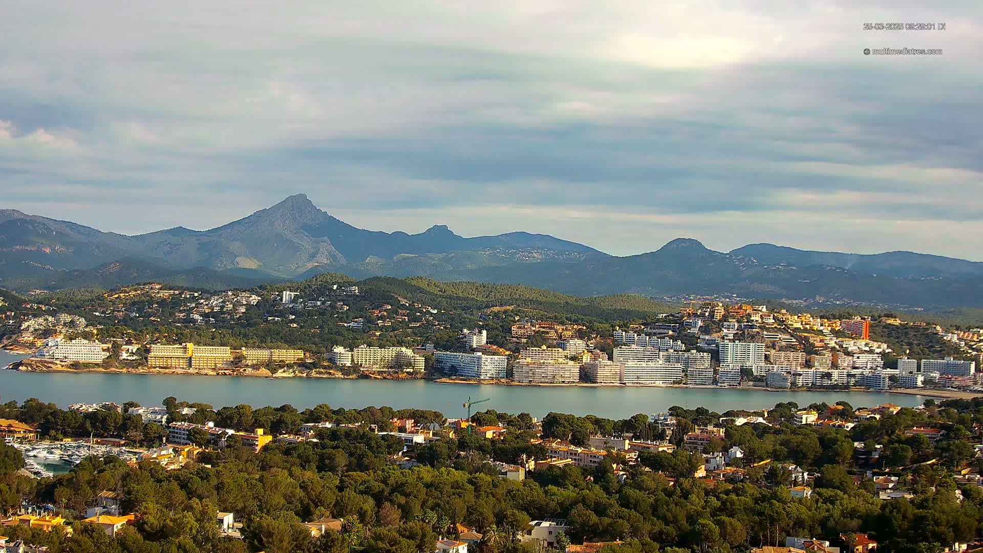 A coastal town nestled between a calm bay and a mountain range under a partly cloudy sky.