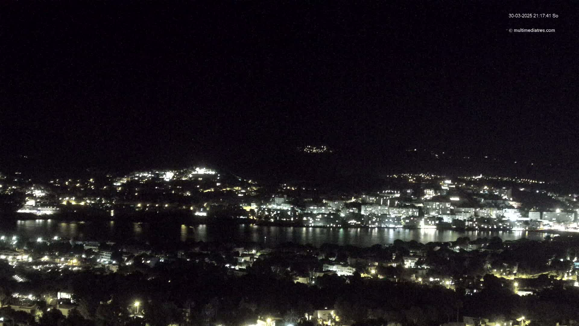A nighttime view of a city illuminated by lights, situated alongside a body of dark water, under a clear, dark sky.