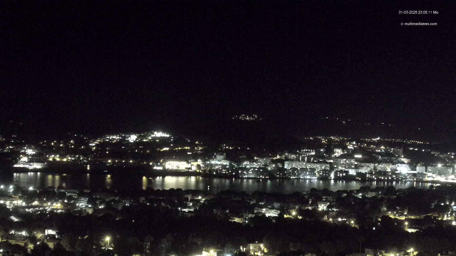 A nighttime view of a city spread across a hillside and along a body of water, under a clear, dark sky.