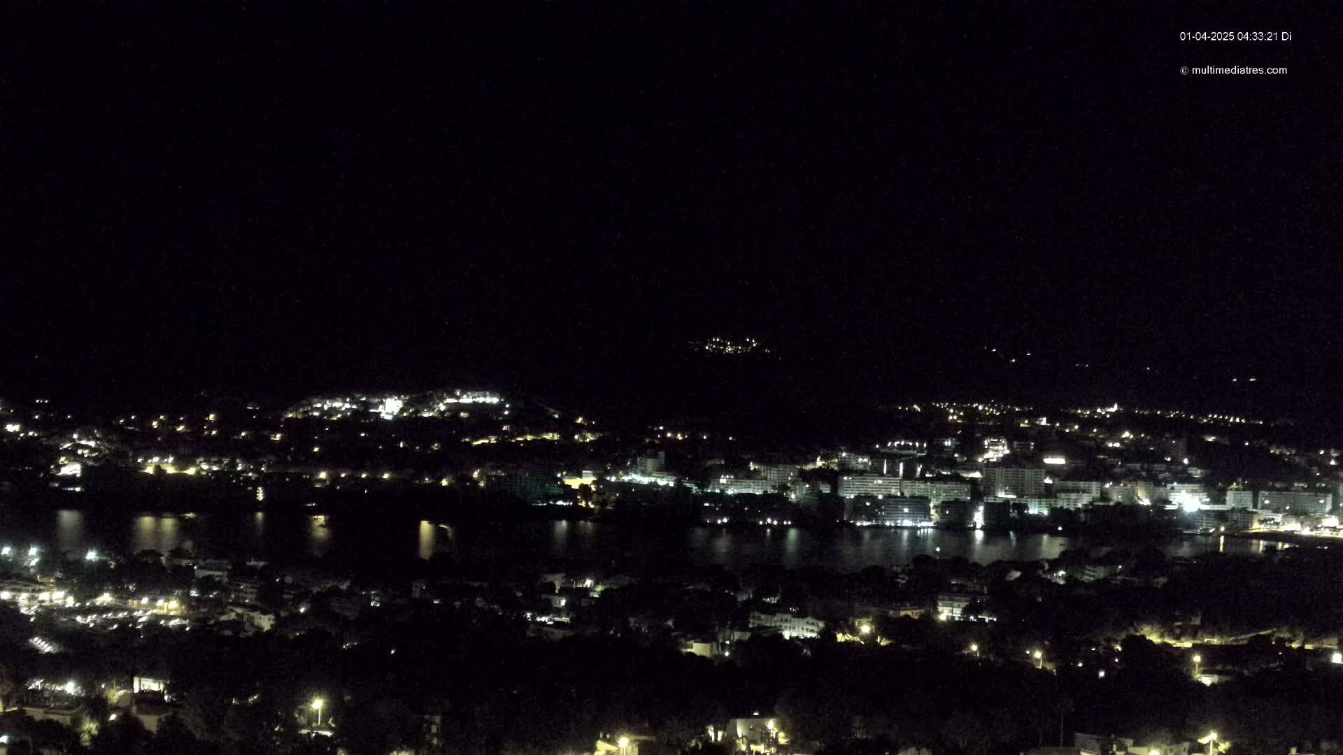 A nighttime view of a city and body of water illuminated by scattered lights under a clear, dark sky.