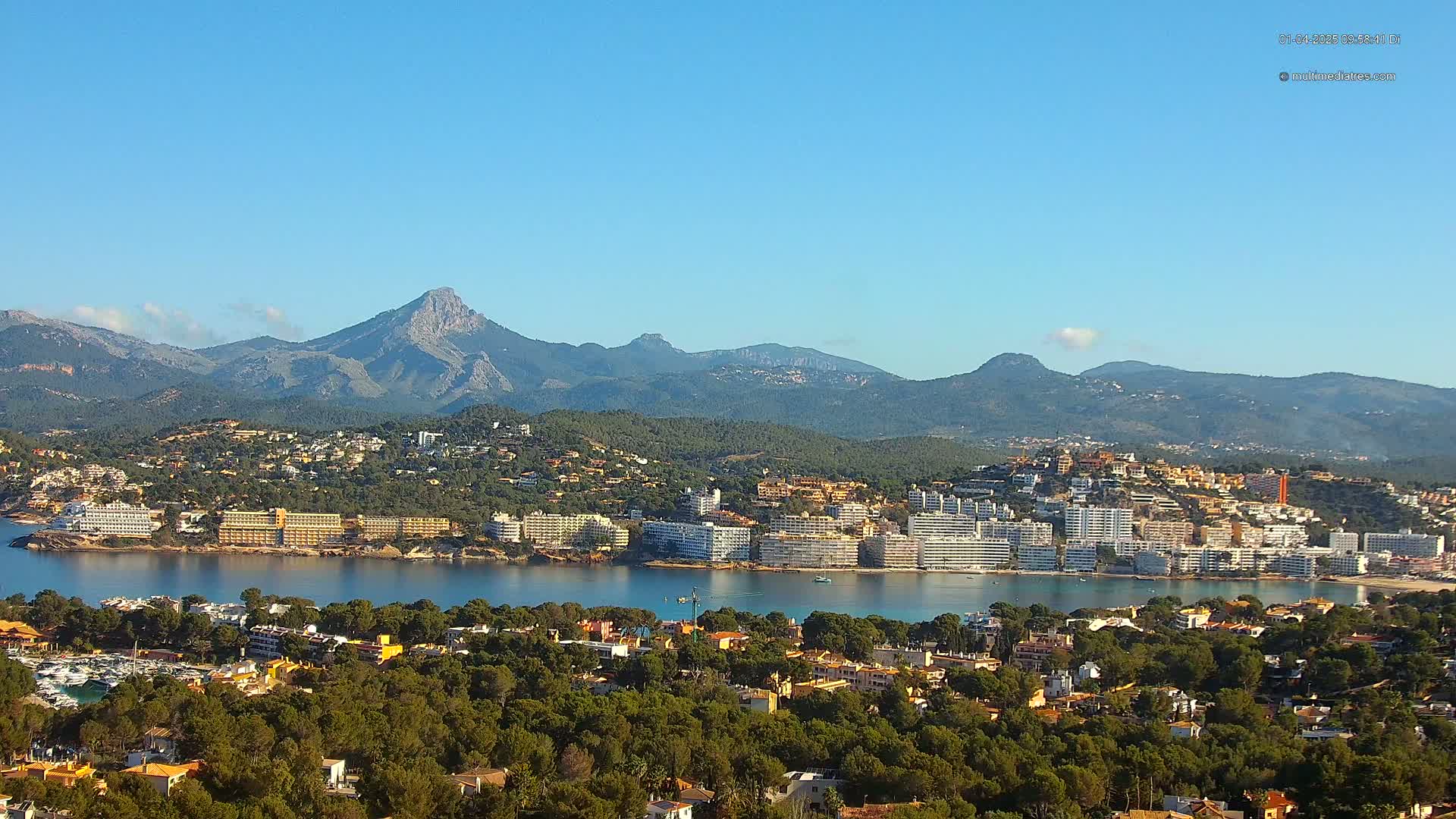 A coastal town with numerous buildings sits nestled at the base of a mountain range under a clear blue sky.
