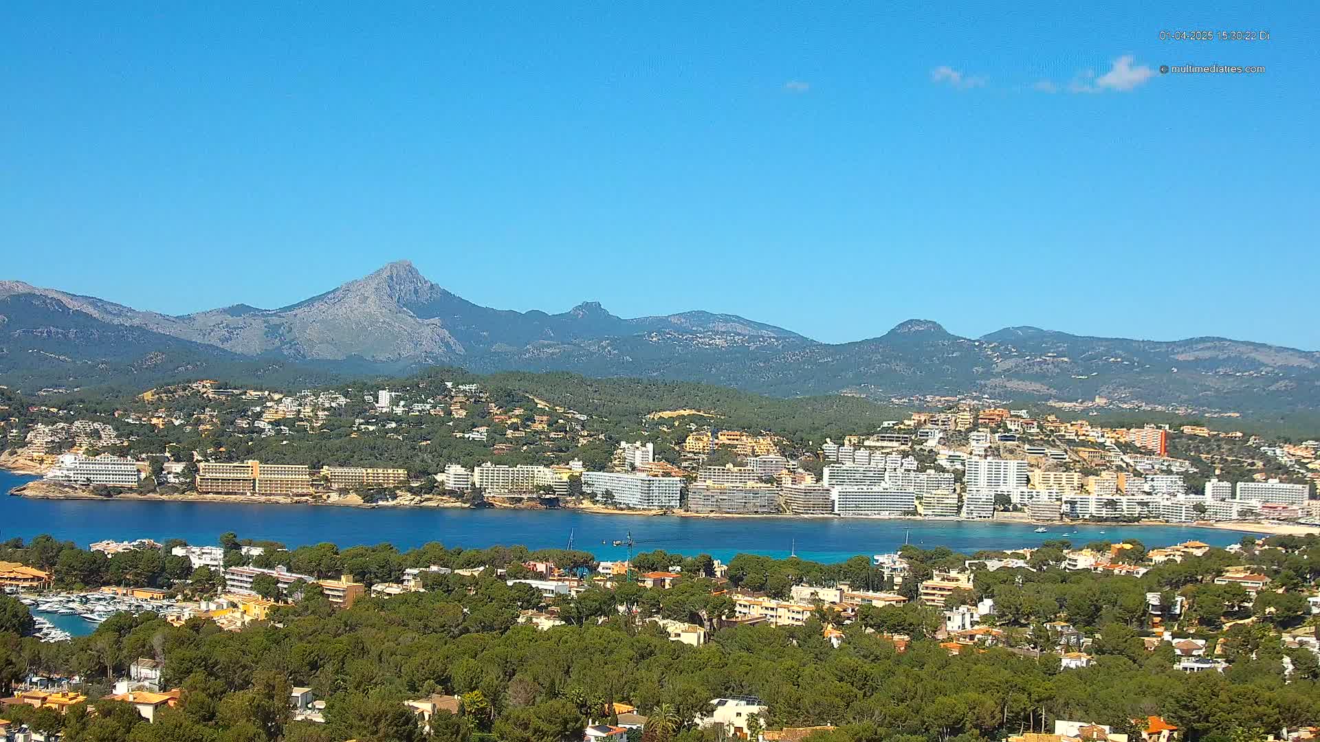 A coastal town nestled amongst green hills and mountains, under a clear blue sky, with a calm bay separating the town from a larger body of water.