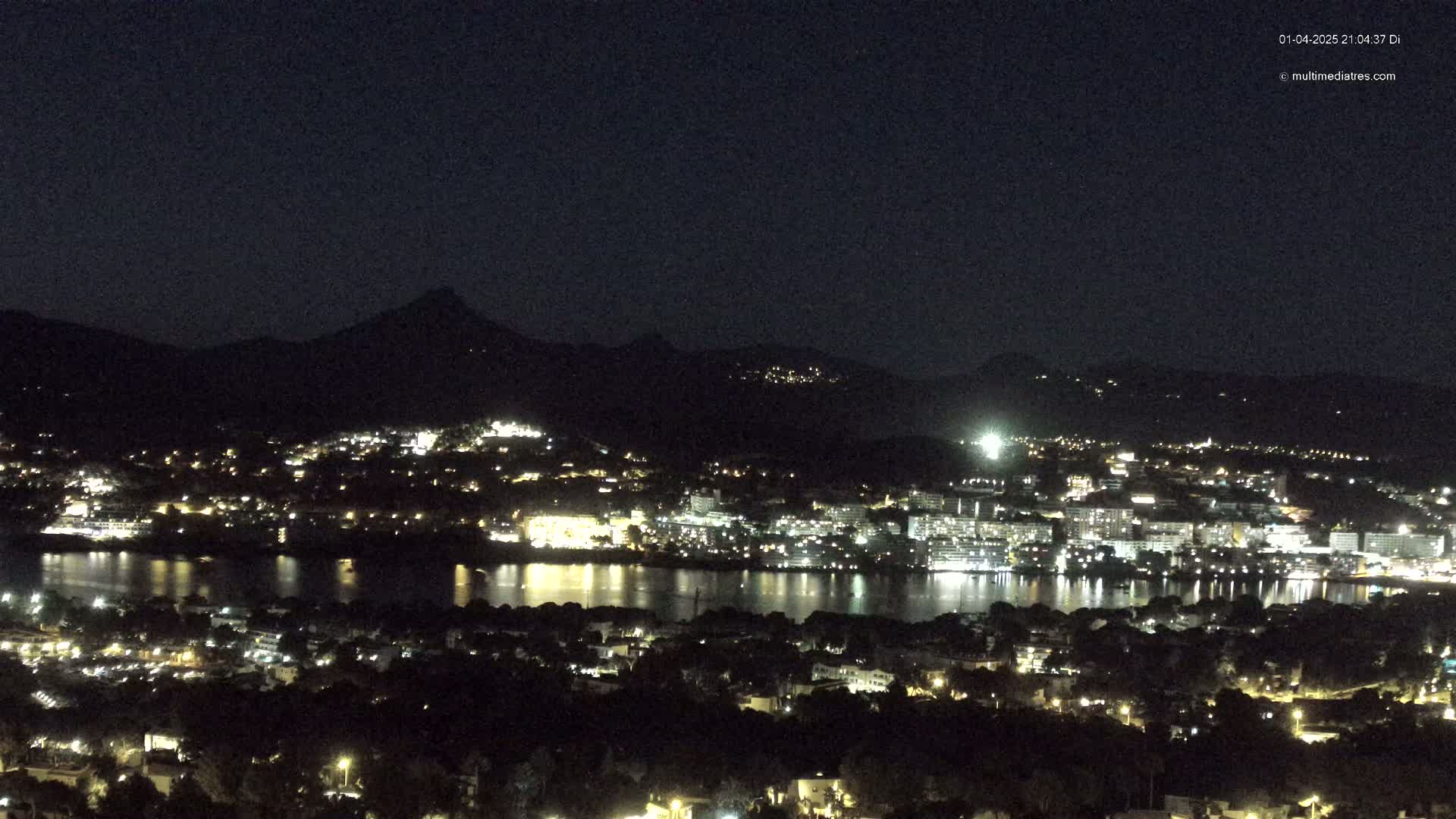 A nighttime view of a city situated on a body of water, nestled between dark hills under a clear sky.