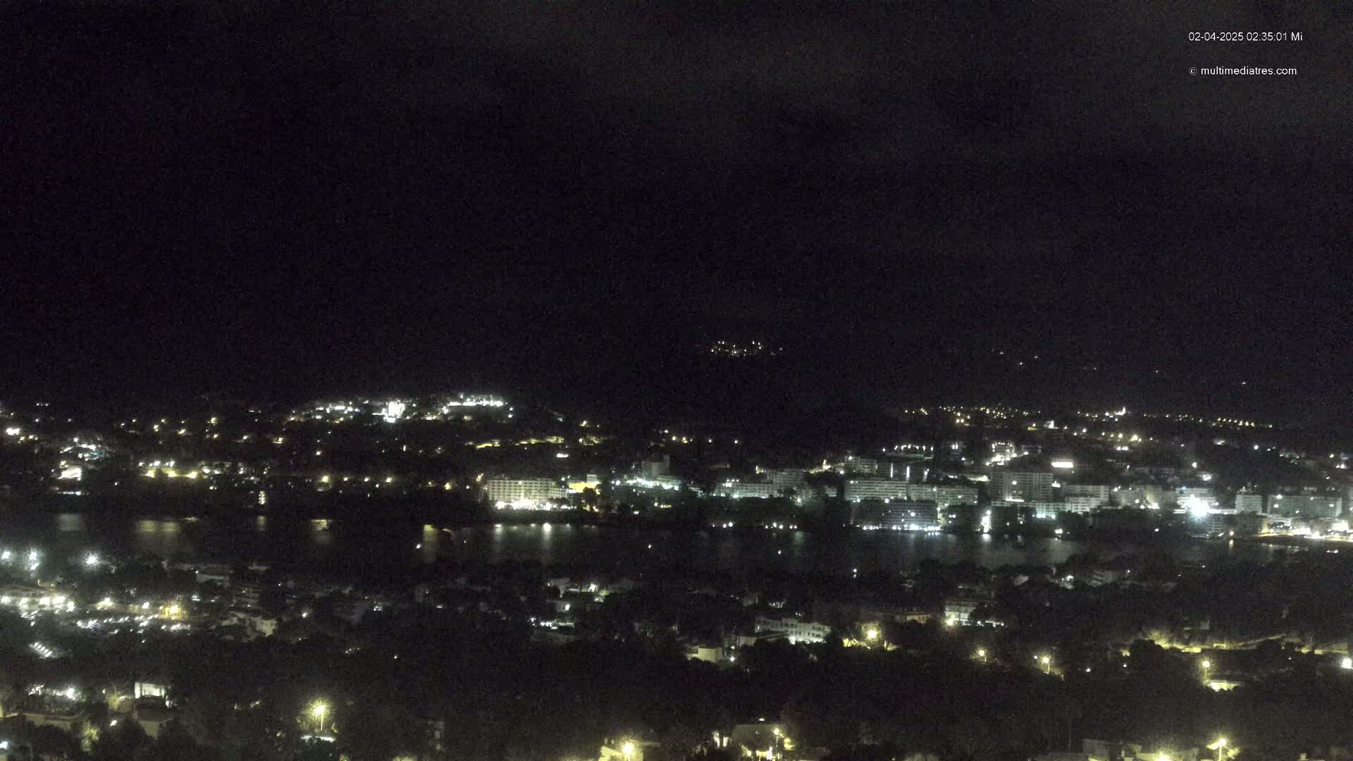 A nighttime view of a coastal city with buildings illuminated along a dark body of water under a cloudy sky.