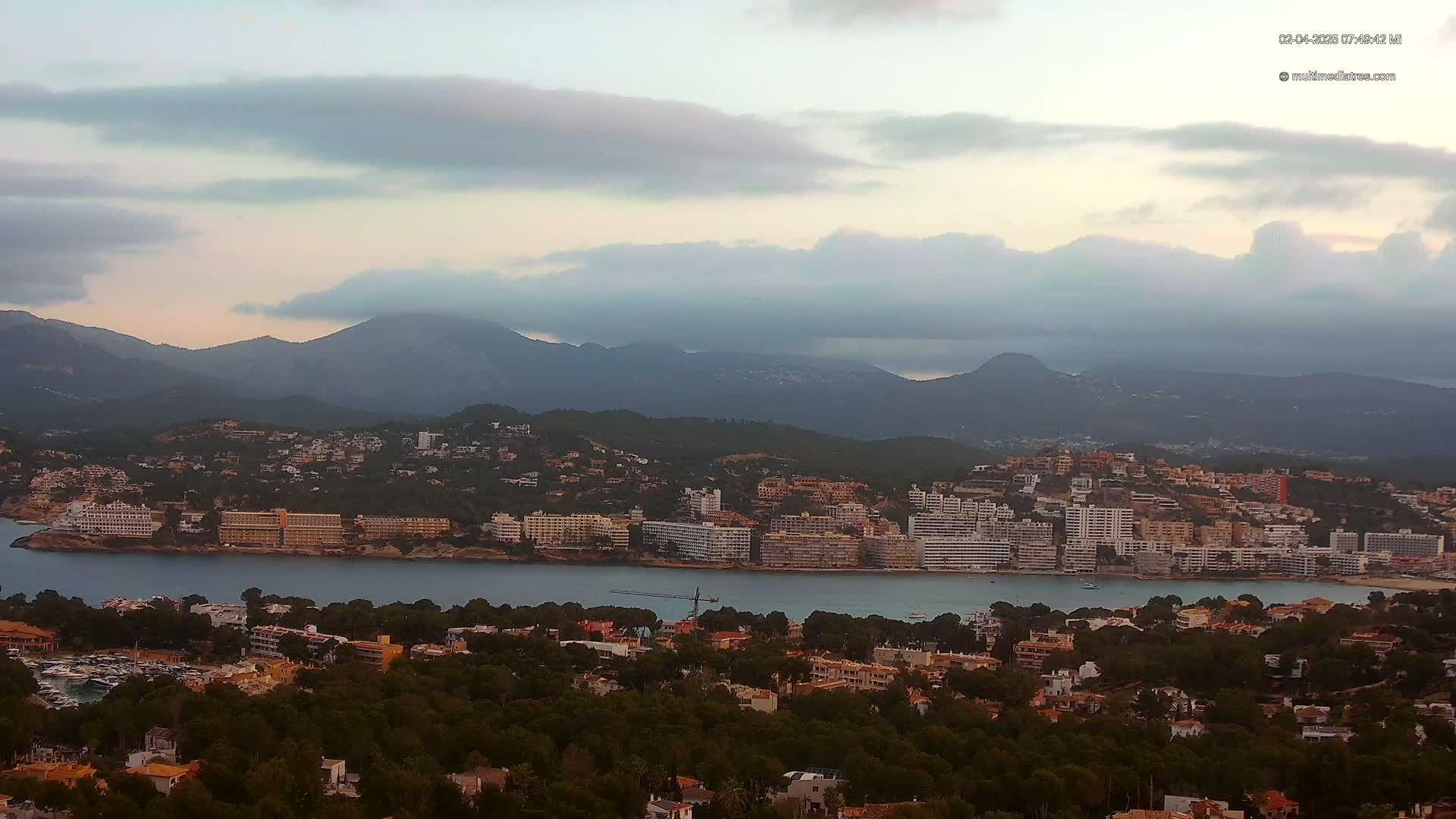 A coastal town nestled between a calm bay and a mountain range under a cloudy sky.