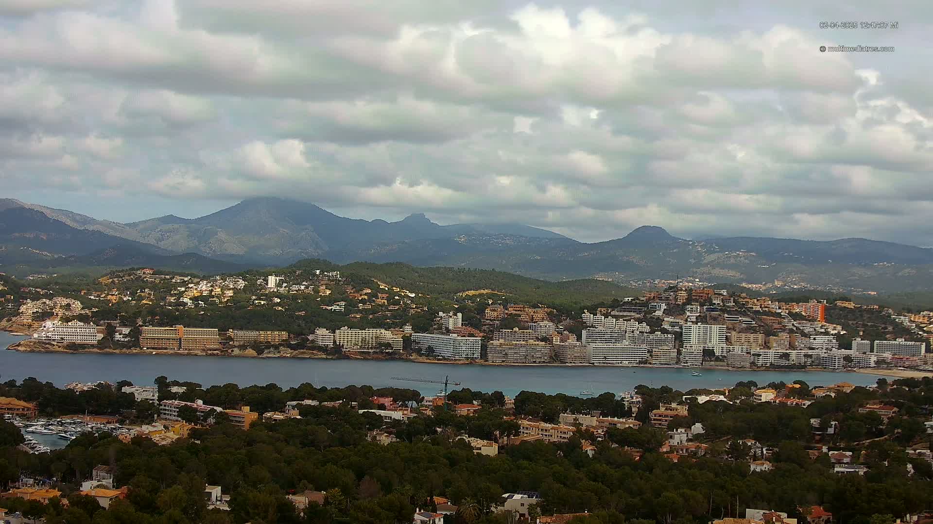 A coastal town with numerous buildings nestled among green hills, overlooks a calm bay under a cloudy sky, with mountains in the background.