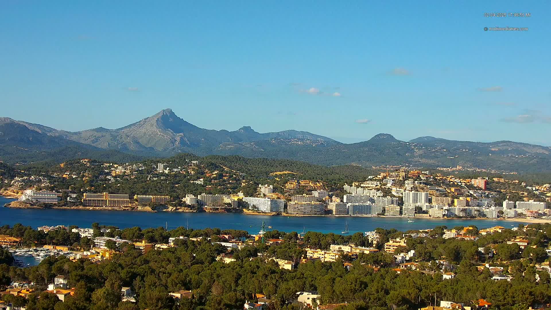 A coastal town nestled between a calm bay and a mountain range under a clear blue sky.