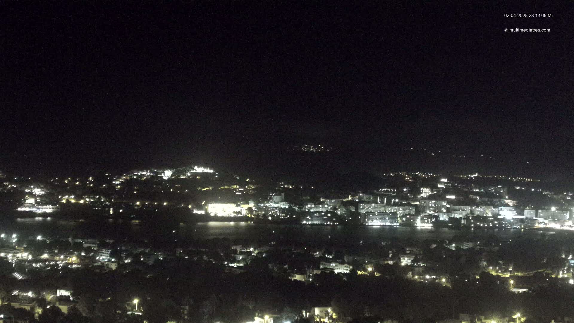 A nighttime, high-angle view shows a coastal city illuminated by lights, with a dark body of water separating a portion of the city from the hilltop vantage point.