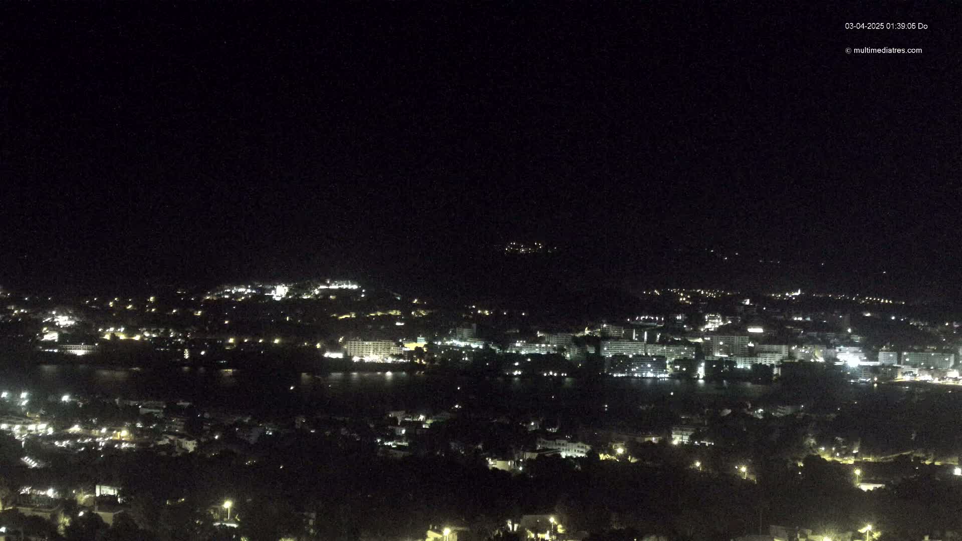 A nighttime, long shot of a coastal town with many lights illuminating buildings and streets, overlooking a dark body of water under a clear, dark sky.