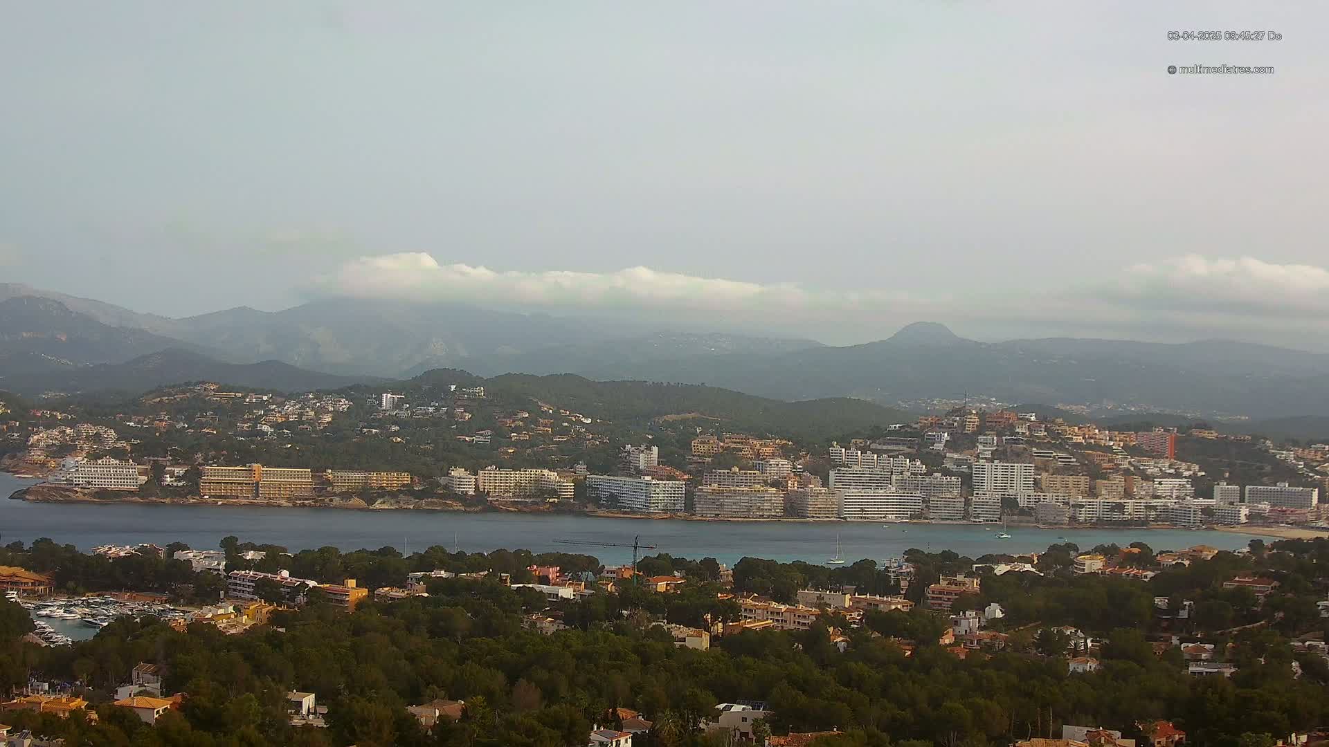 A coastal town nestled between a body of water and a mountain range under a hazy sky.