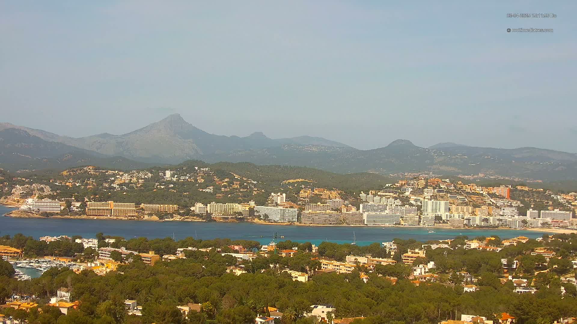 A coastal town nestled amongst green hills and mountains under a hazy sky overlooks a calm bay.