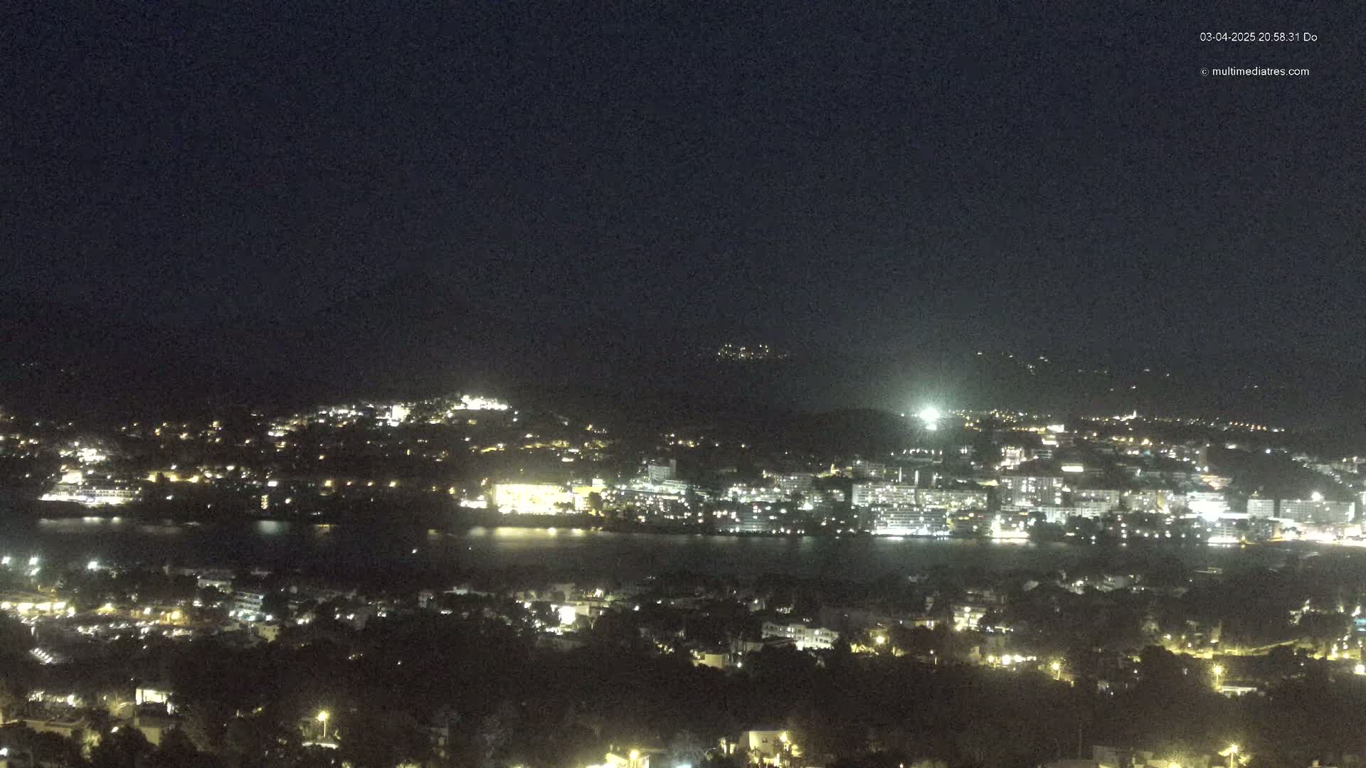 A nighttime, long-range view of a city sprawling across a body of water, with hills visible in the background under a clear, dark sky.