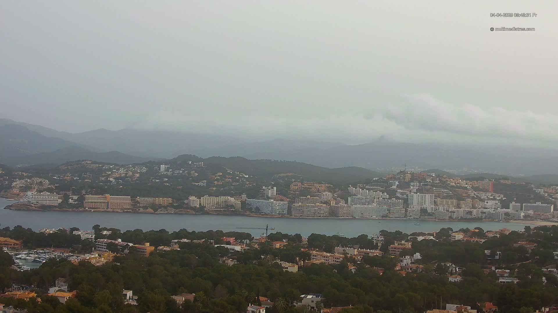 A coastal town nestled between a calm bay and hazy mountains under an overcast sky.