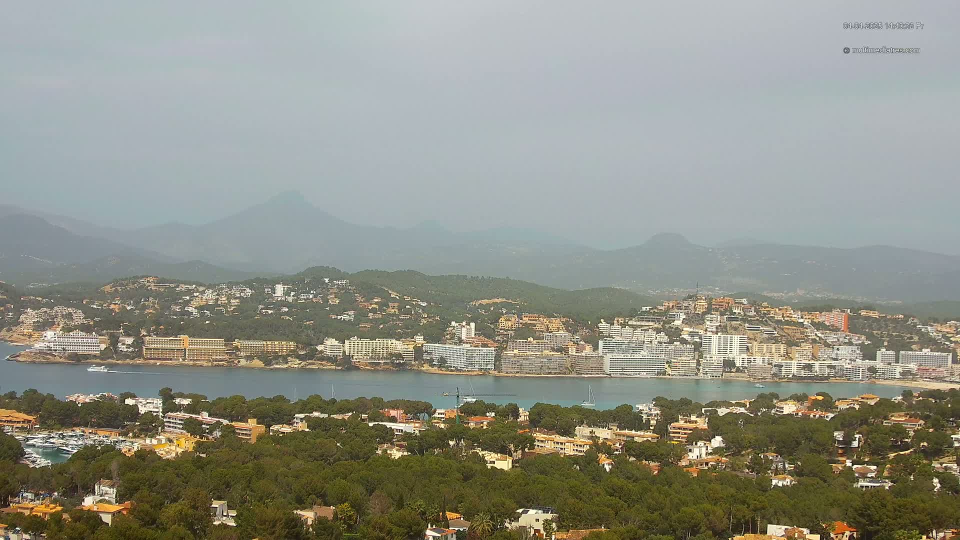 A hazy coastal town with numerous buildings and hotels nestled between a calm bay and a mountain range.