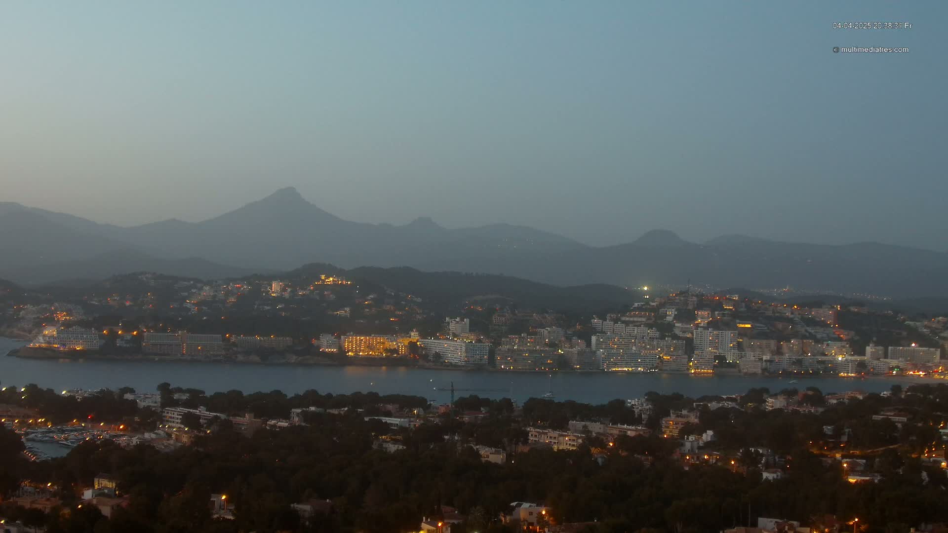 A coastal town with illuminated buildings at dusk, nestled between a calm body of water and hazy mountains under a clear, twilight sky.