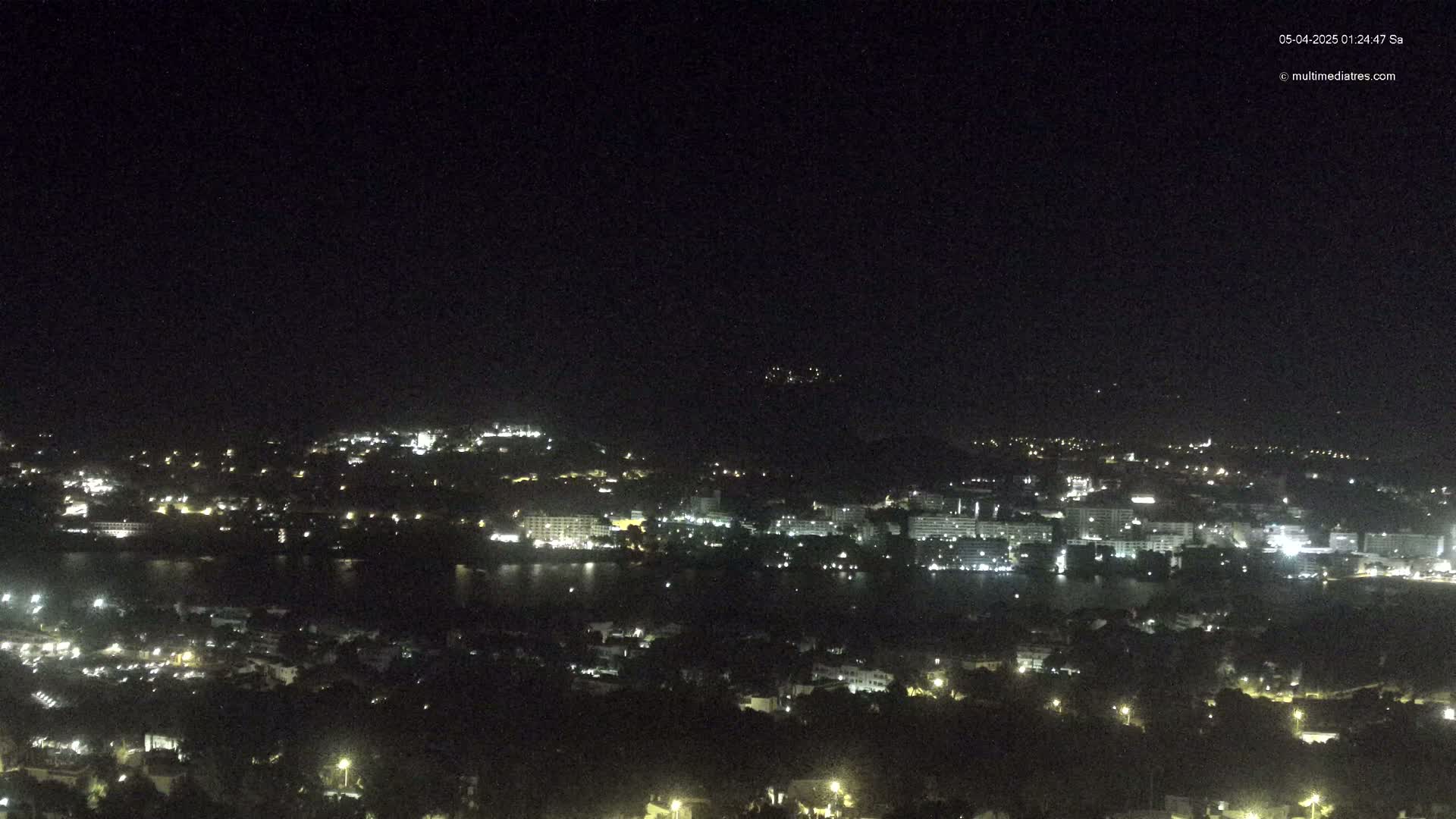 A nighttime, aerial view shows a coastal town with scattered lights along a dark body of water, under a clear, dark sky.