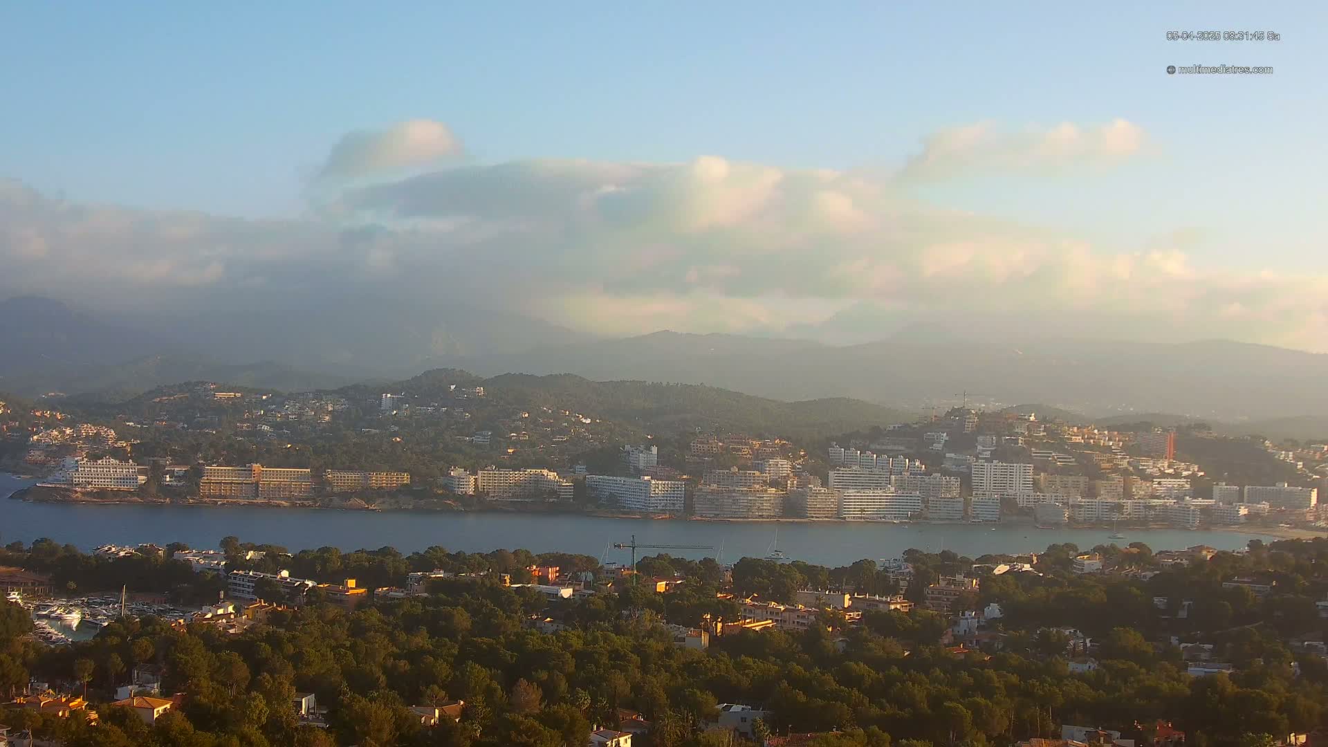 A coastal town with numerous buildings sits nestled between a calm bay and hazy mountains under a partly cloudy sky.