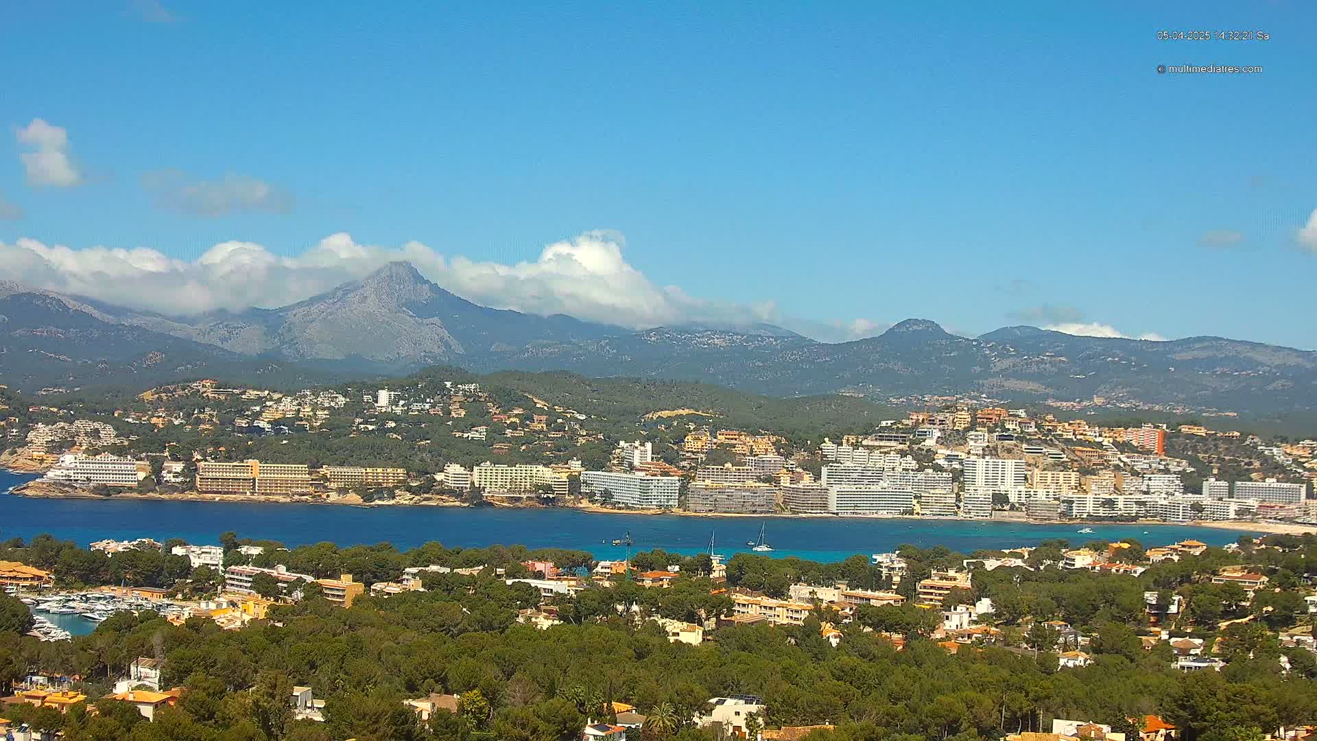 A coastal town with hotels and residential buildings nestled along a bay, backed by mountains partially covered in clouds under a mostly clear blue sky.