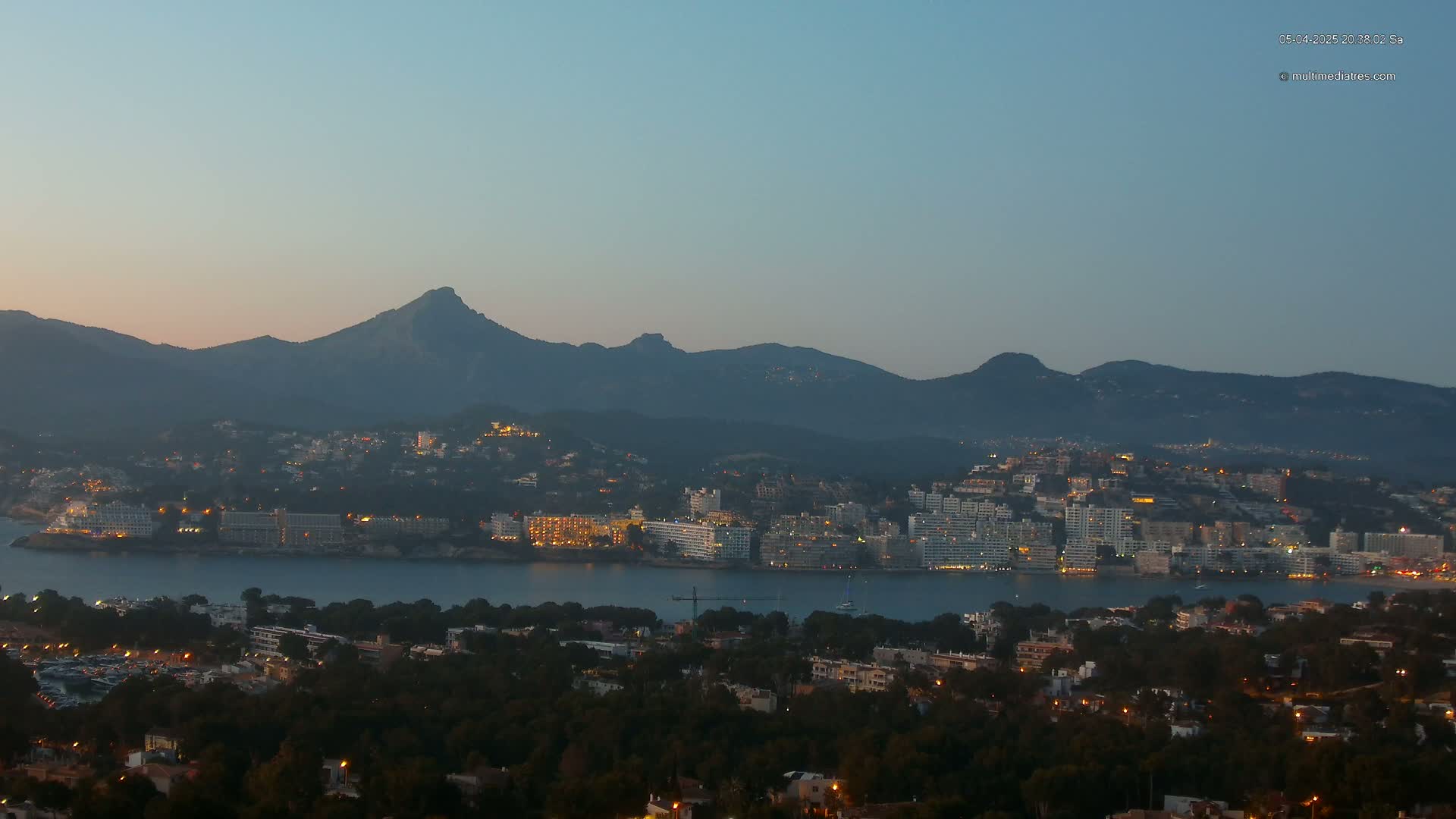 A coastal city with many lights at twilight, nestled at the base of a dark mountain range under a clear, dusky sky.
