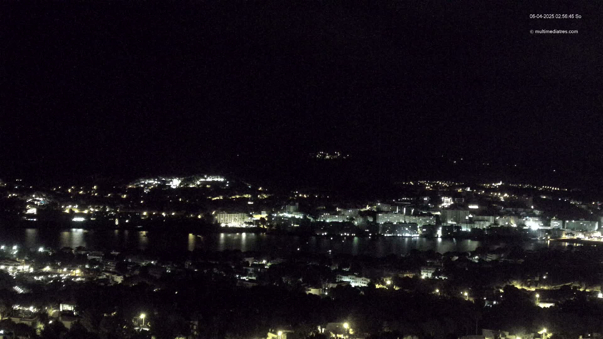 A nighttime, high-angle view shows a coastal town with buildings and lights reflecting on a dark body of water under a clear, dark sky.
