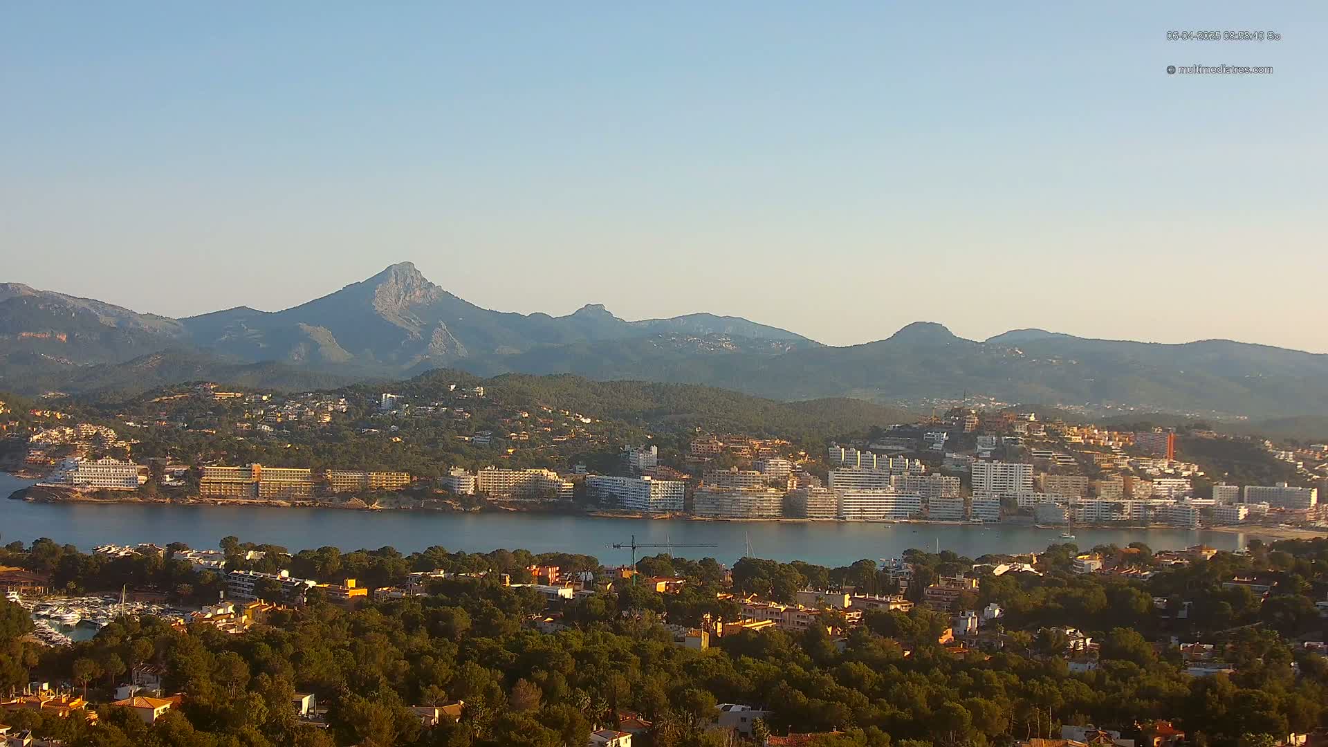 A coastal town nestled between a calm bay and a mountain range under a clear, sunny sky.