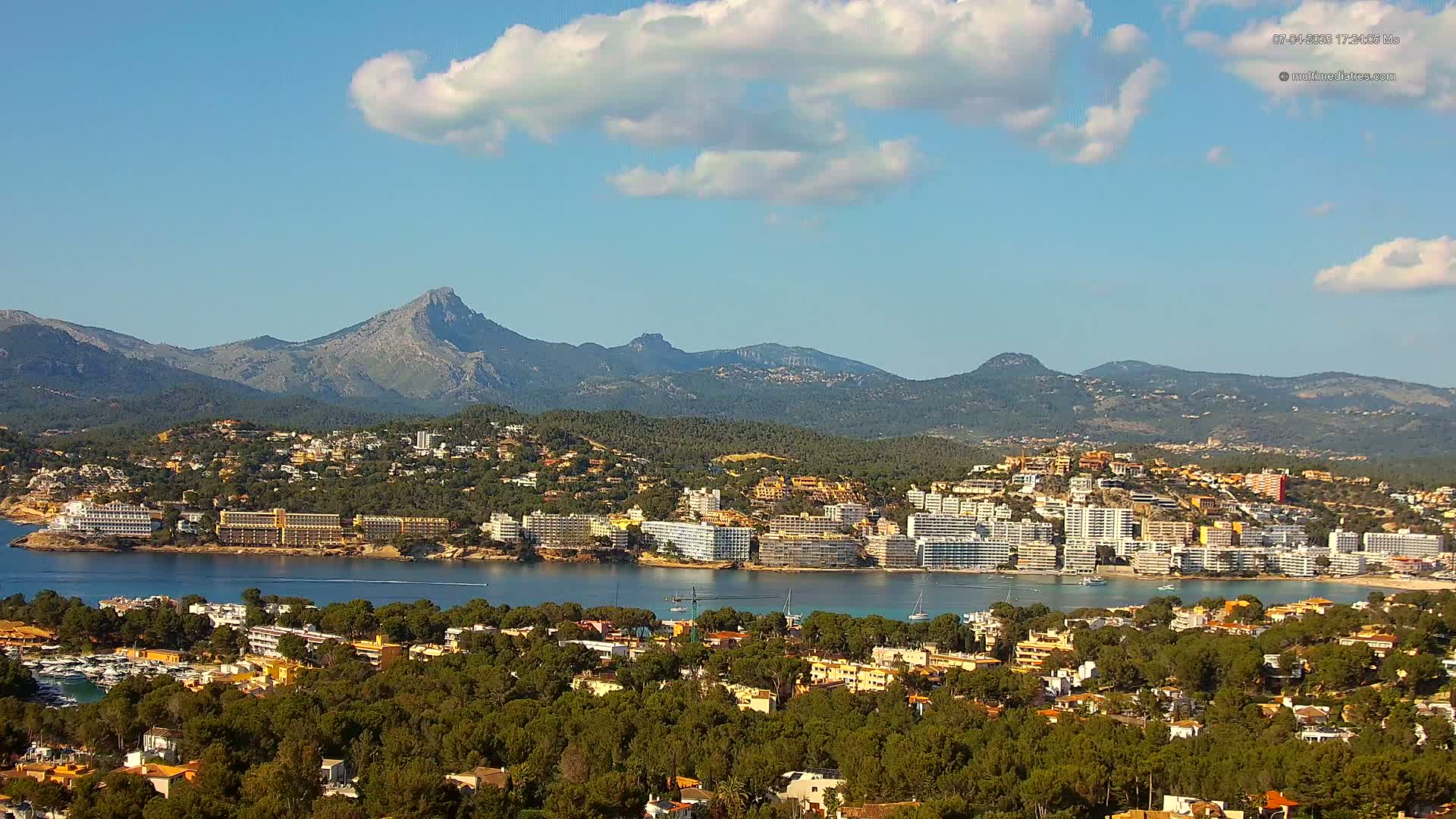 A coastal town nestled among green hills and mountains, under a mostly sunny sky with a few scattered clouds, overlooks a calm bay.