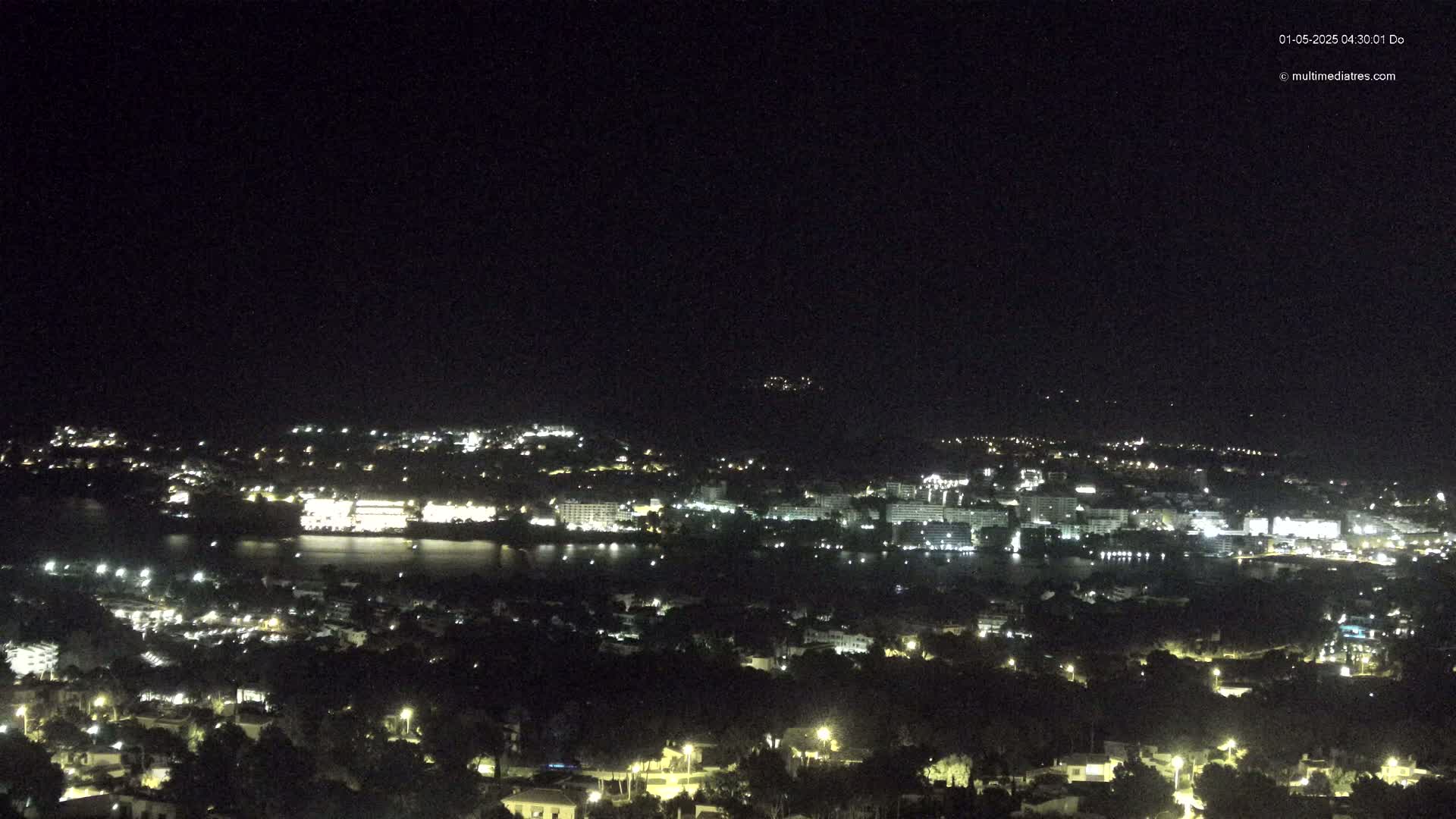 A nighttime view of a coastal town with many lights illuminating buildings and streets, and a dark body of water separating a hilltop area from the main town.