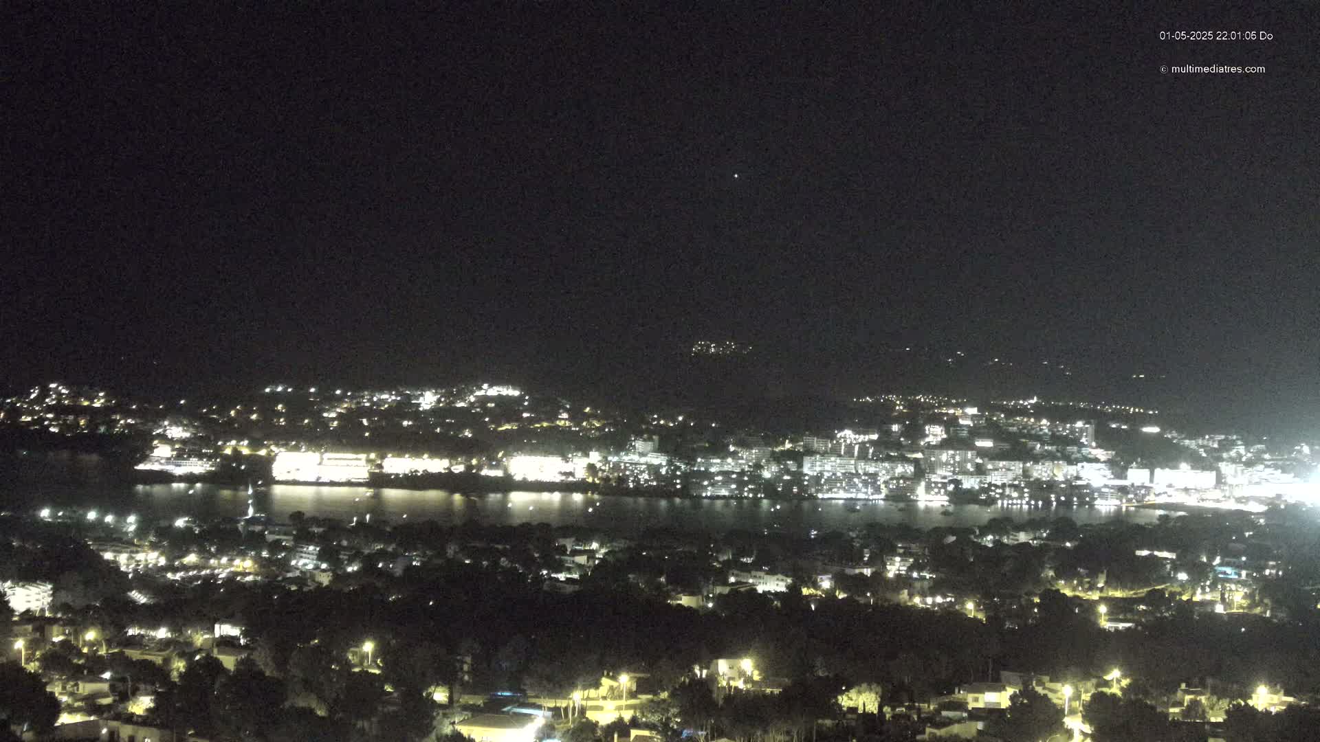 A nighttime, aerial view shows a coastal town illuminated with many lights, situated next to a dark body of water, with hills in the background under a clear sky.