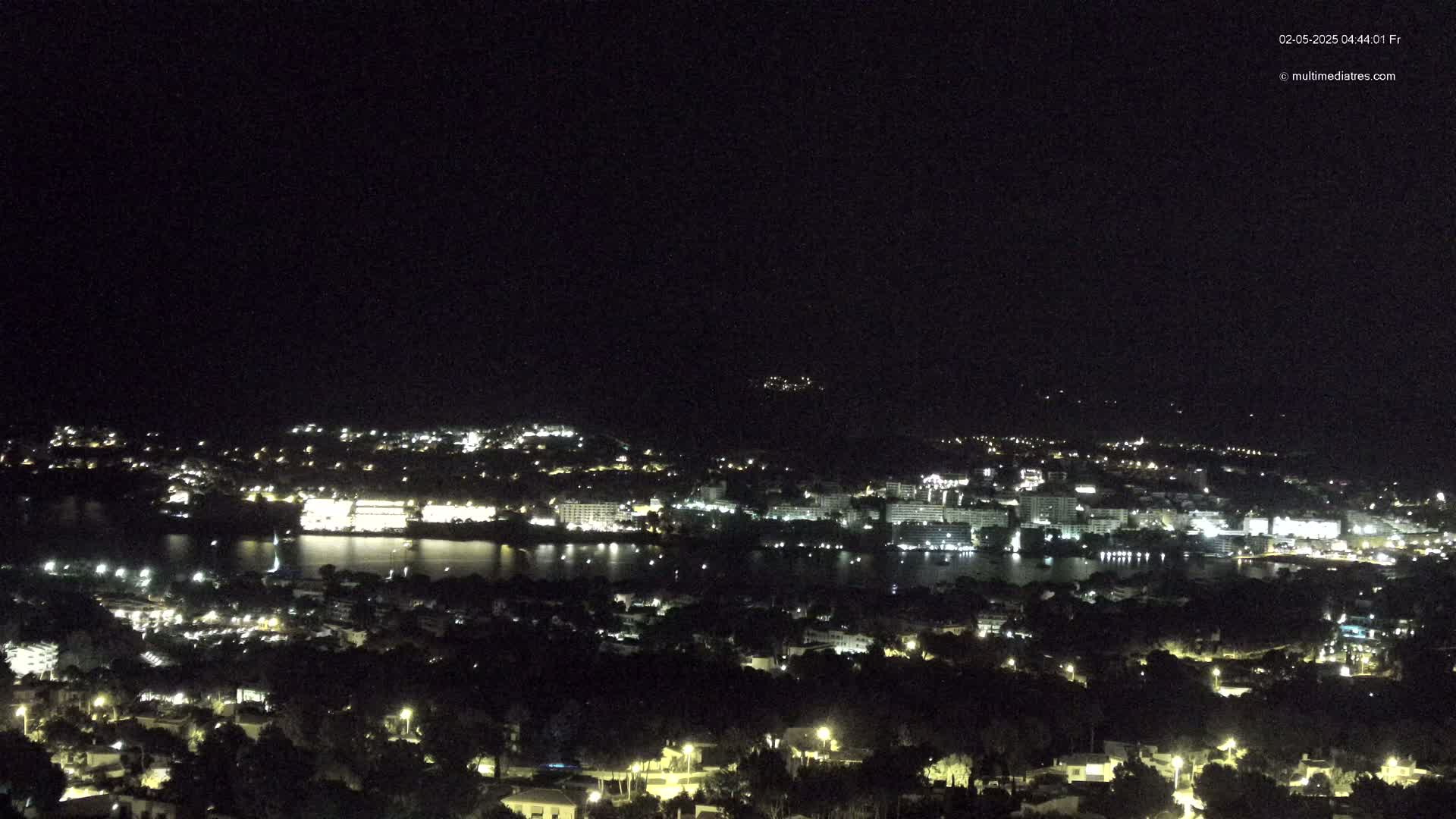 A nighttime, high-angle view shows a coastal town with numerous lights illuminating buildings and streets along a dark body of water, under a clear, dark sky.