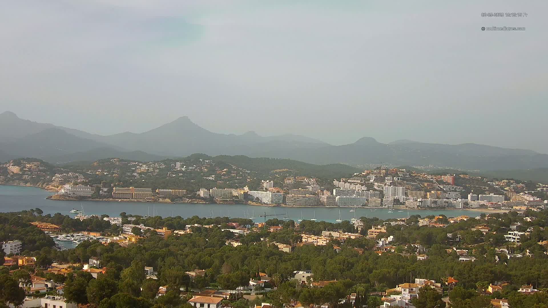 A hazy coastal town nestled among green hills and a calm bay, backed by a mountain range.