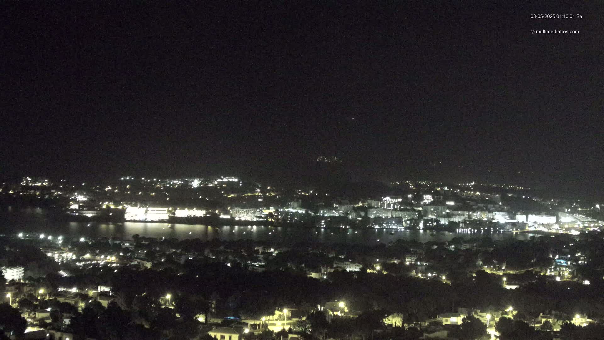 A nighttime, long-range view of a coastal town with many lights illuminating buildings and streets, situated next to a calm body of water under a clear, dark sky.