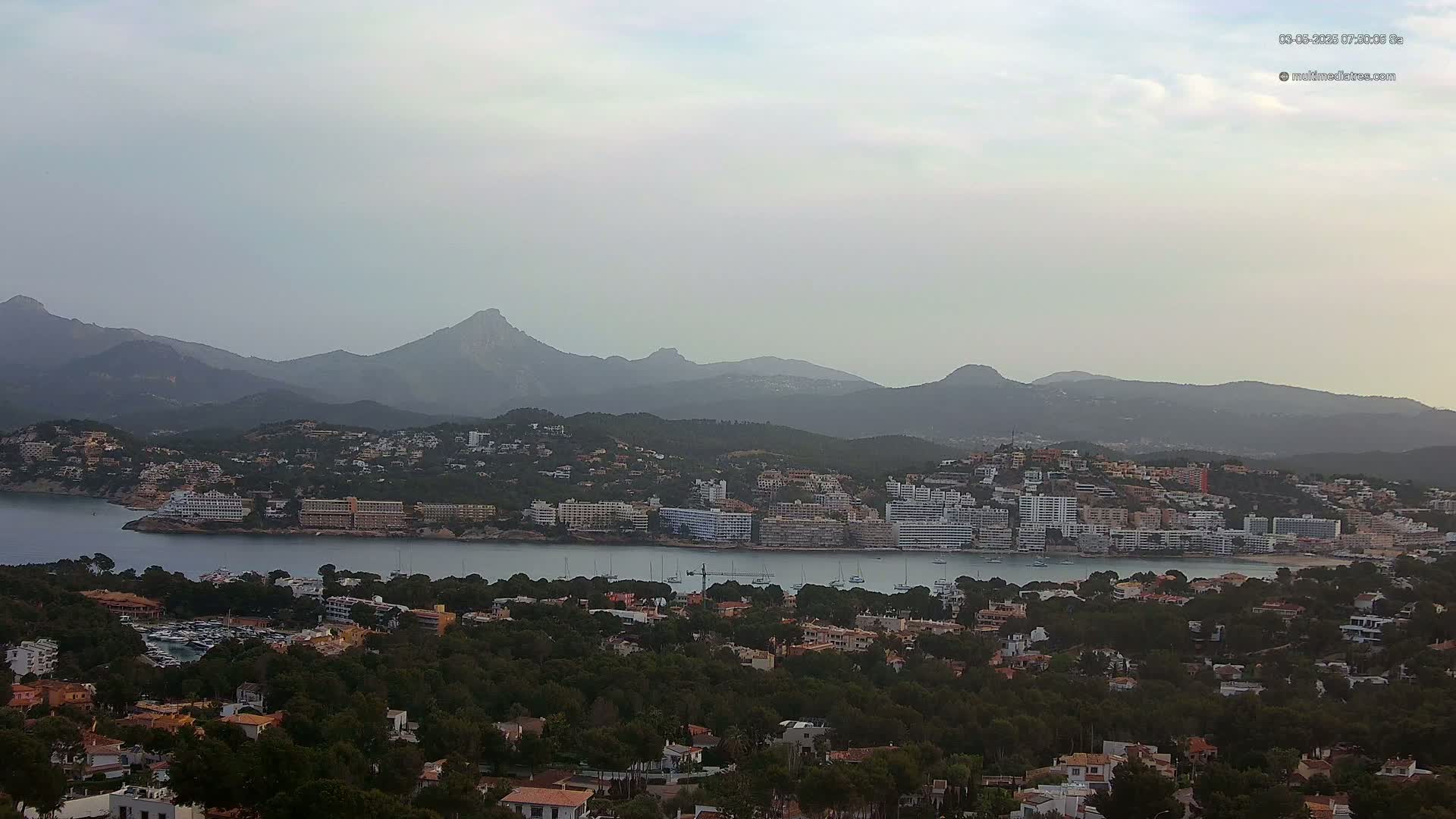 A coastal town nestled between a calm bay and hazy mountains under a cloudy sky.