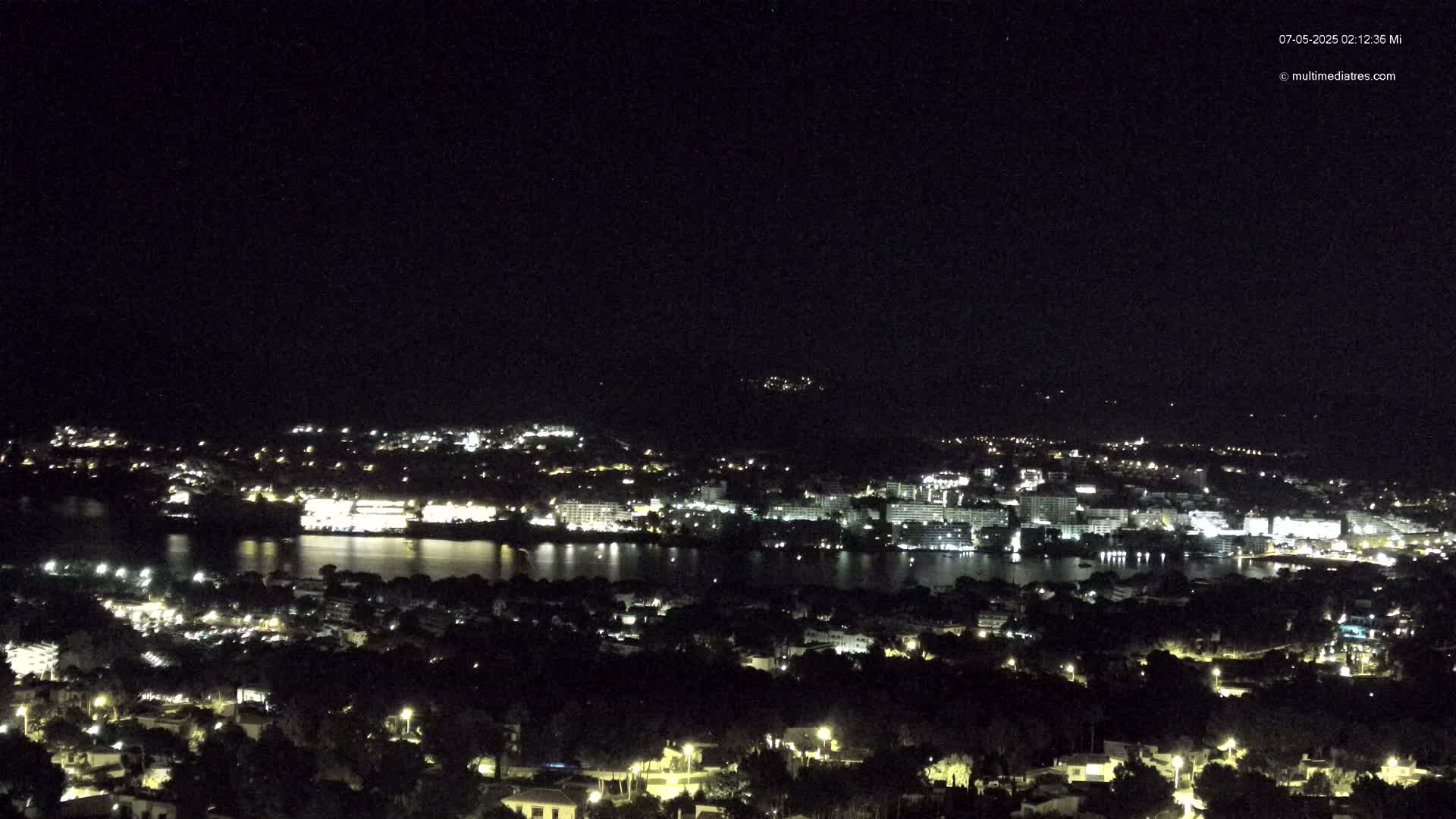 A nighttime, high-angle view shows a body of water reflecting the lights of a city nestled along its shores, with additional lights illuminating the surrounding hills and landscape.