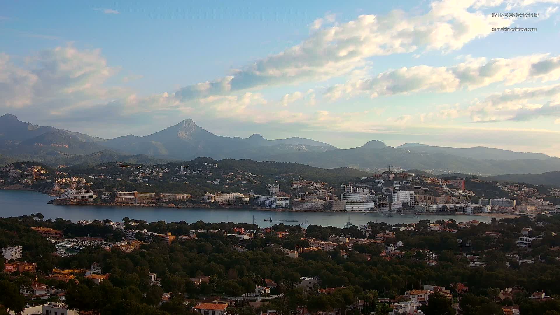 A coastal town nestled among green hills and mountains under a partly cloudy sky, with a body of water separating the town from a larger mountainous area.